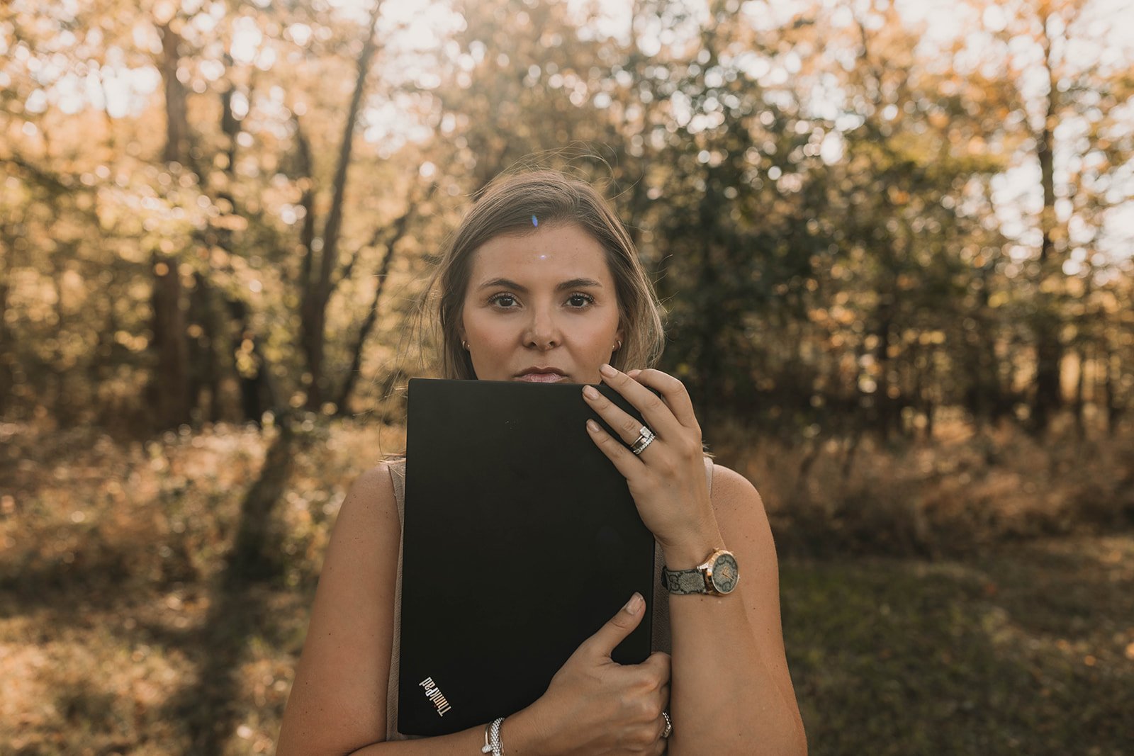 Kerri M. Roberts holding a black tablet in front of her face in an outdoor setting with autumn trees and leaves.