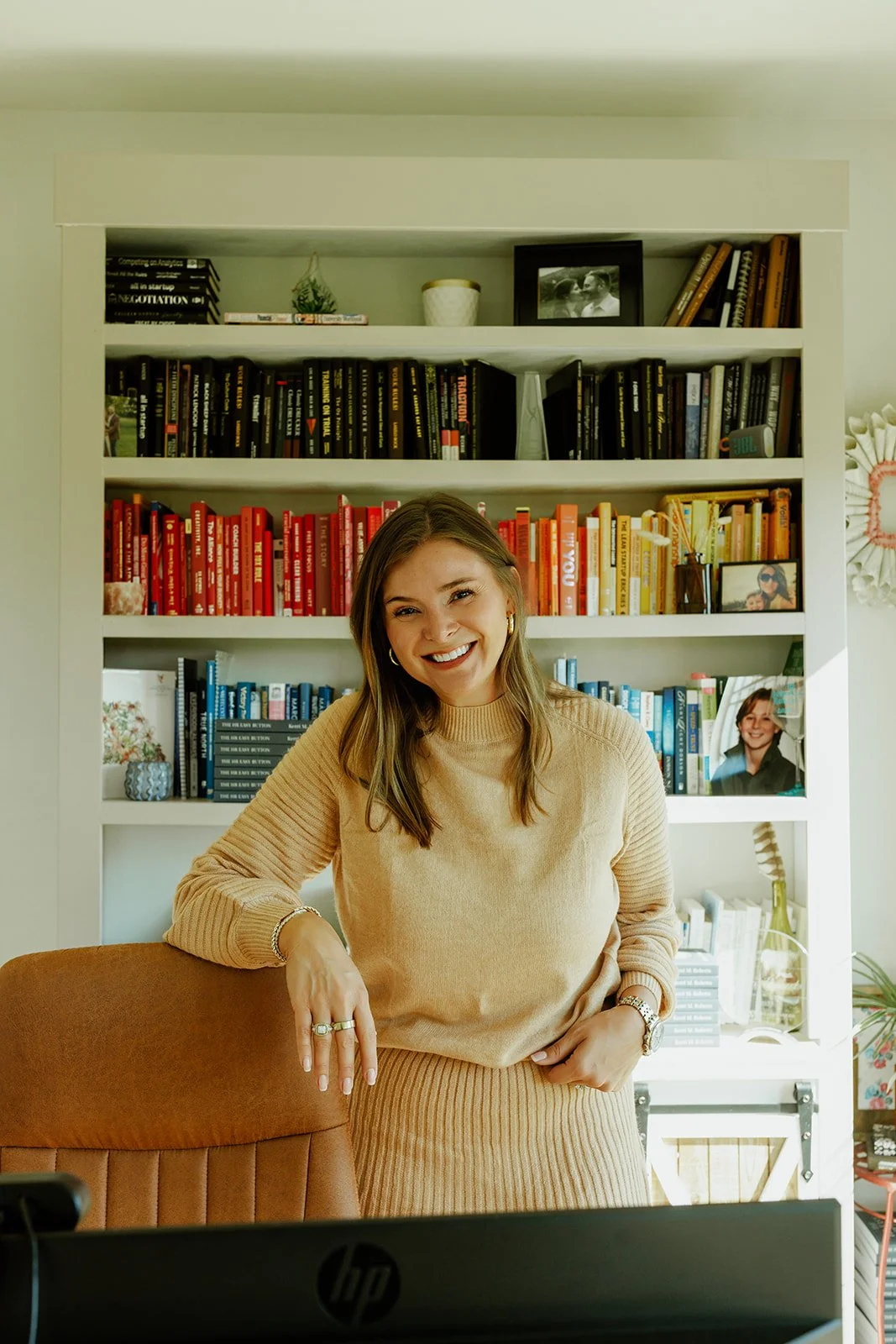 Kerri M. Roberts smiling and leaning on a chair in front of a bookshelf with a rainbow of books and framed photos.