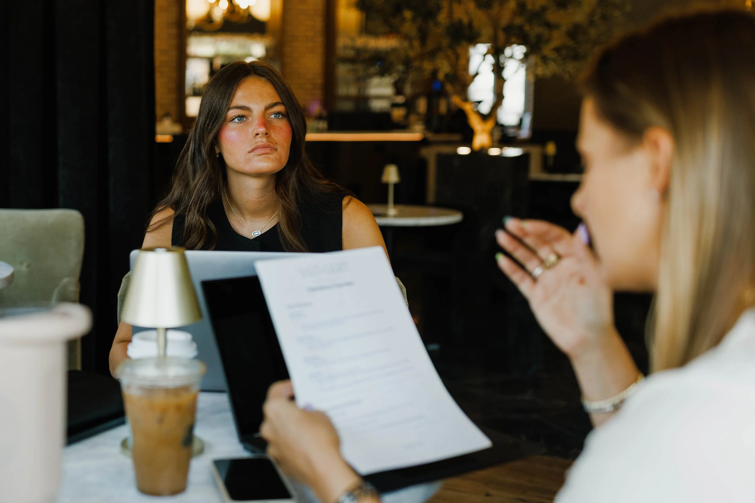 Two women are sitting at a table in a cafe. One woman with long dark hair appears upset or serious, looking at the other woman with blonde hair, who is holding a document. There is a drink with ice and a straw in front of the blonde woman.