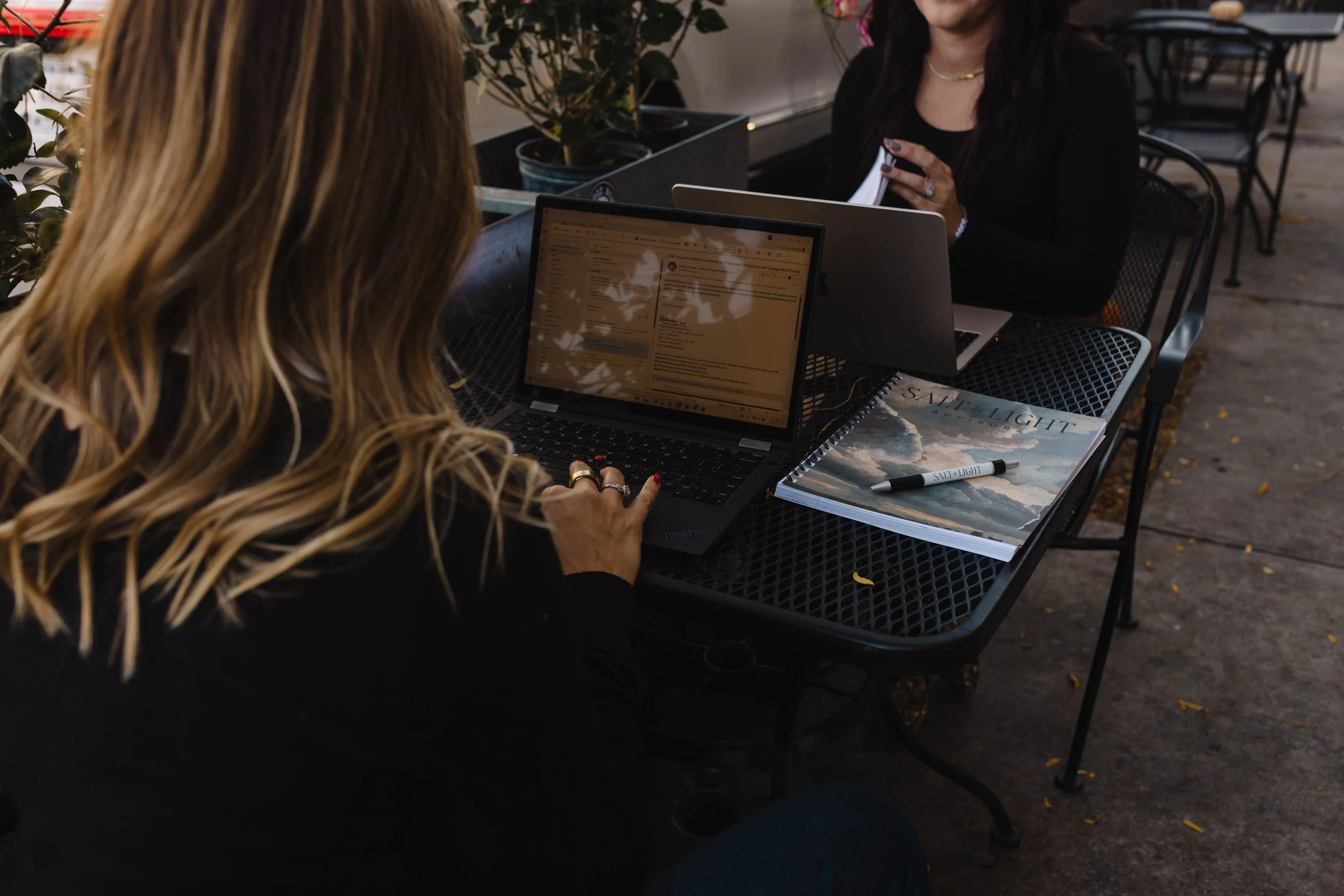 Two women sitting at a black outdoor table using laptops, one with long blonde hair, and a notebook titled 'Salt & Light' with a pen on top of it.