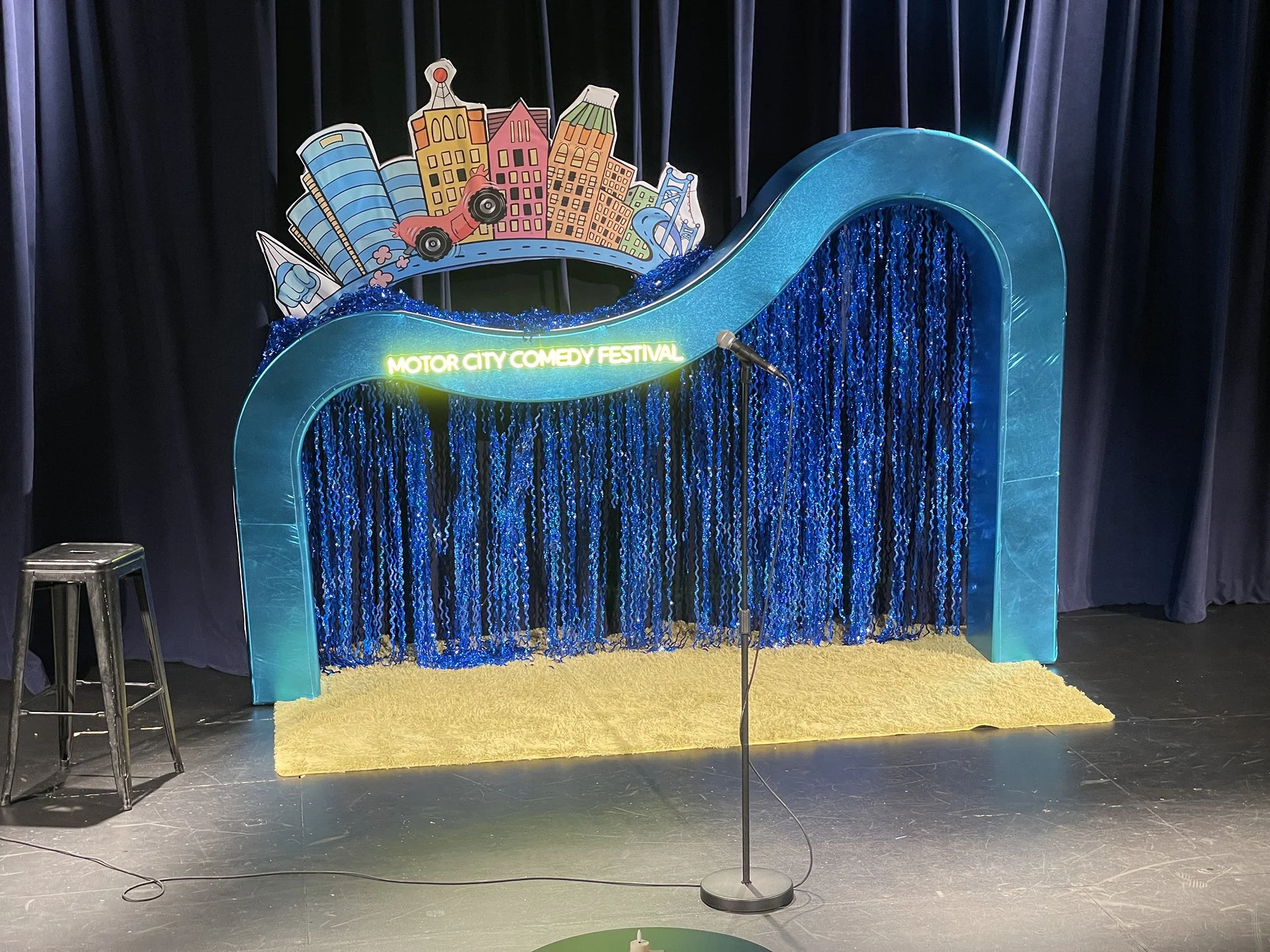 Stage with a blue arch backdrop and a yellow carpet, featuring a sign that reads 'Motor City Comedy Festival,' and a microphone on a stand in the center, with a stool on the left.