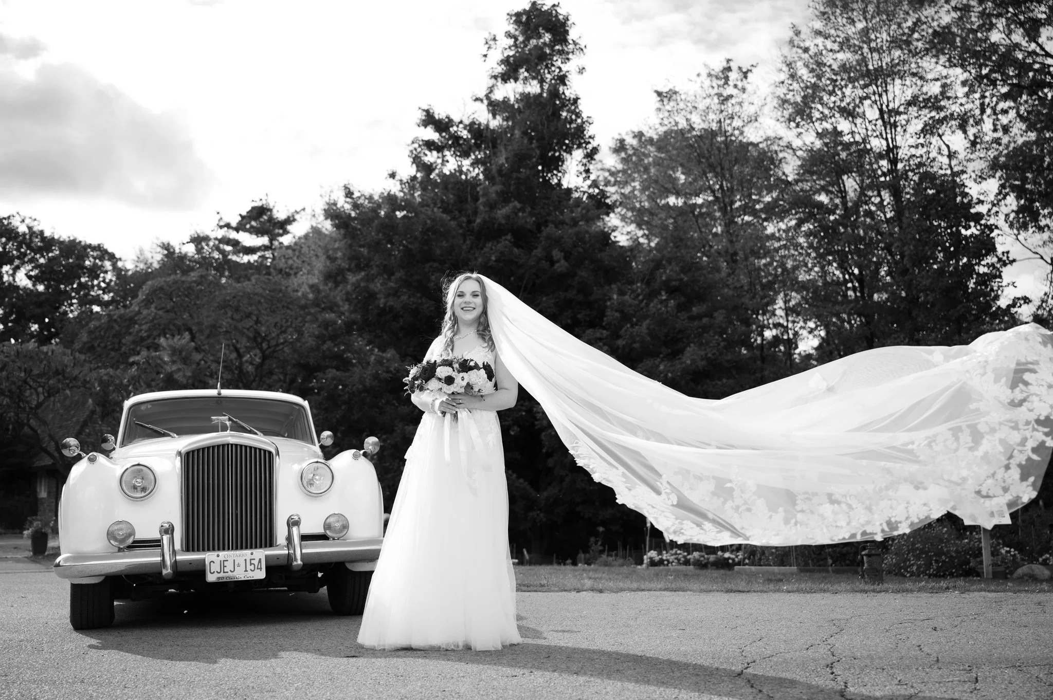 Bride posing with a long flowing veil beside a vintage white car at Riverwood Park in Mississauga.