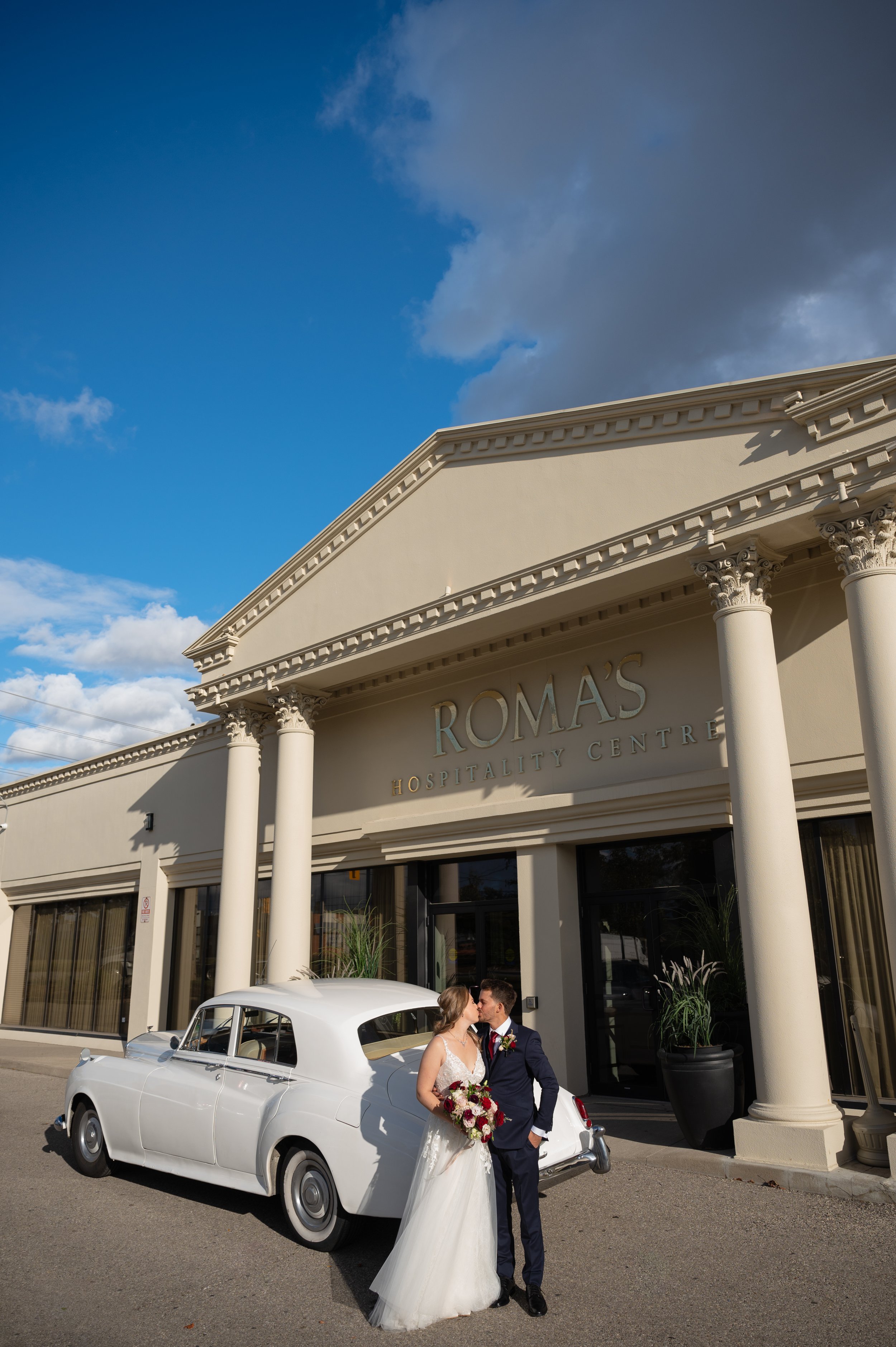 Bride and groom sharing a kiss outside Roma’s Event Space with a vintage white car.
