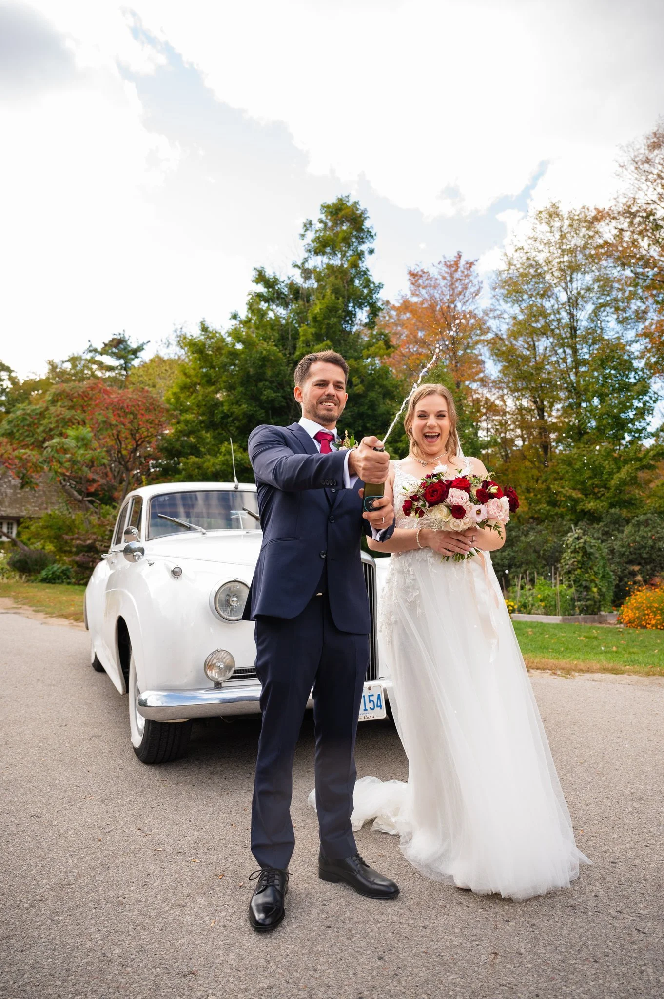 Bride and groom popping a champagne bottle and laughing together during their wedding portraits at Riverwood Park in Mississauga.