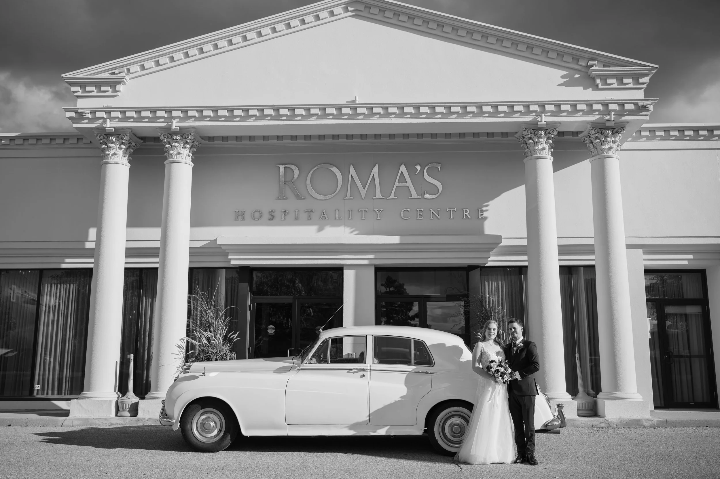Bride and groom standing beside a vintage white car outside Roma’s Hospitality Centre in Mississauga.