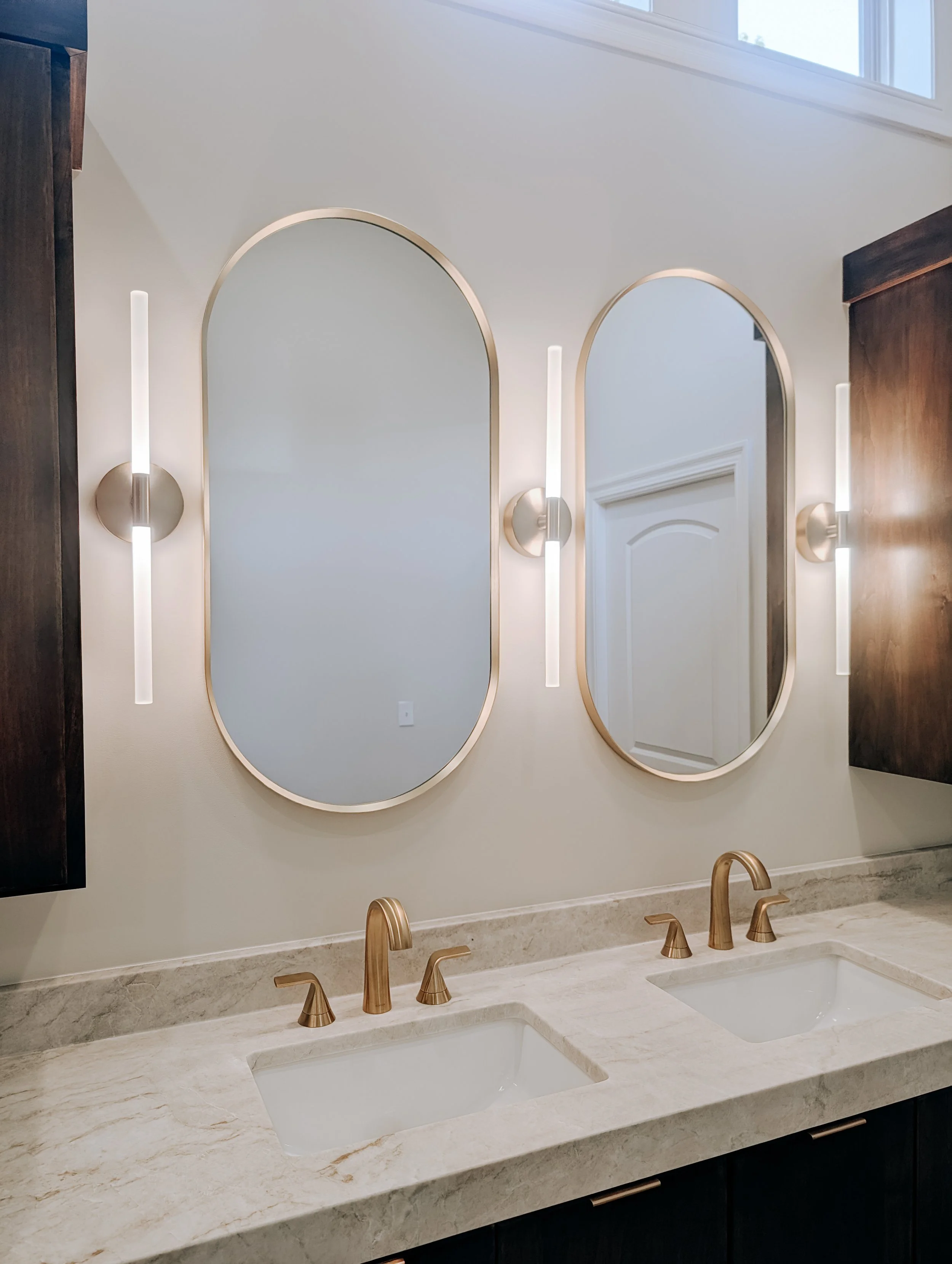 Brushed gold faucets, oval mirrors and vertical vanity sconces above custom double vanity