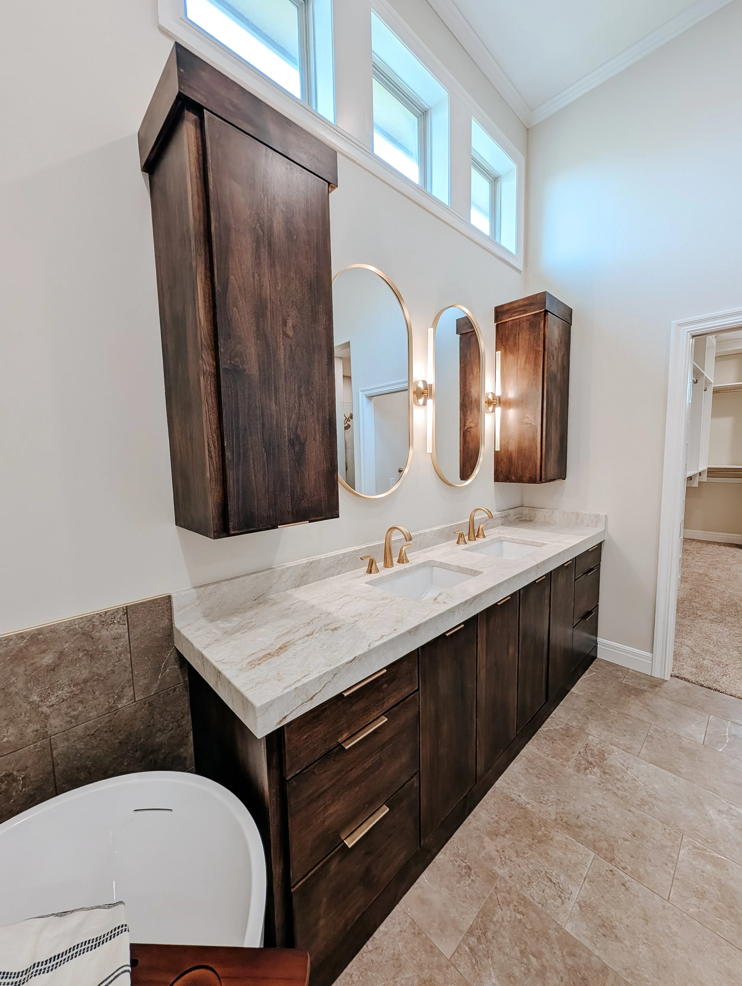 Neutral Lees Summit Bathroom Remodel with dark brown cabinets, a double vanity, gold finishes, sconces and oval mirrors
