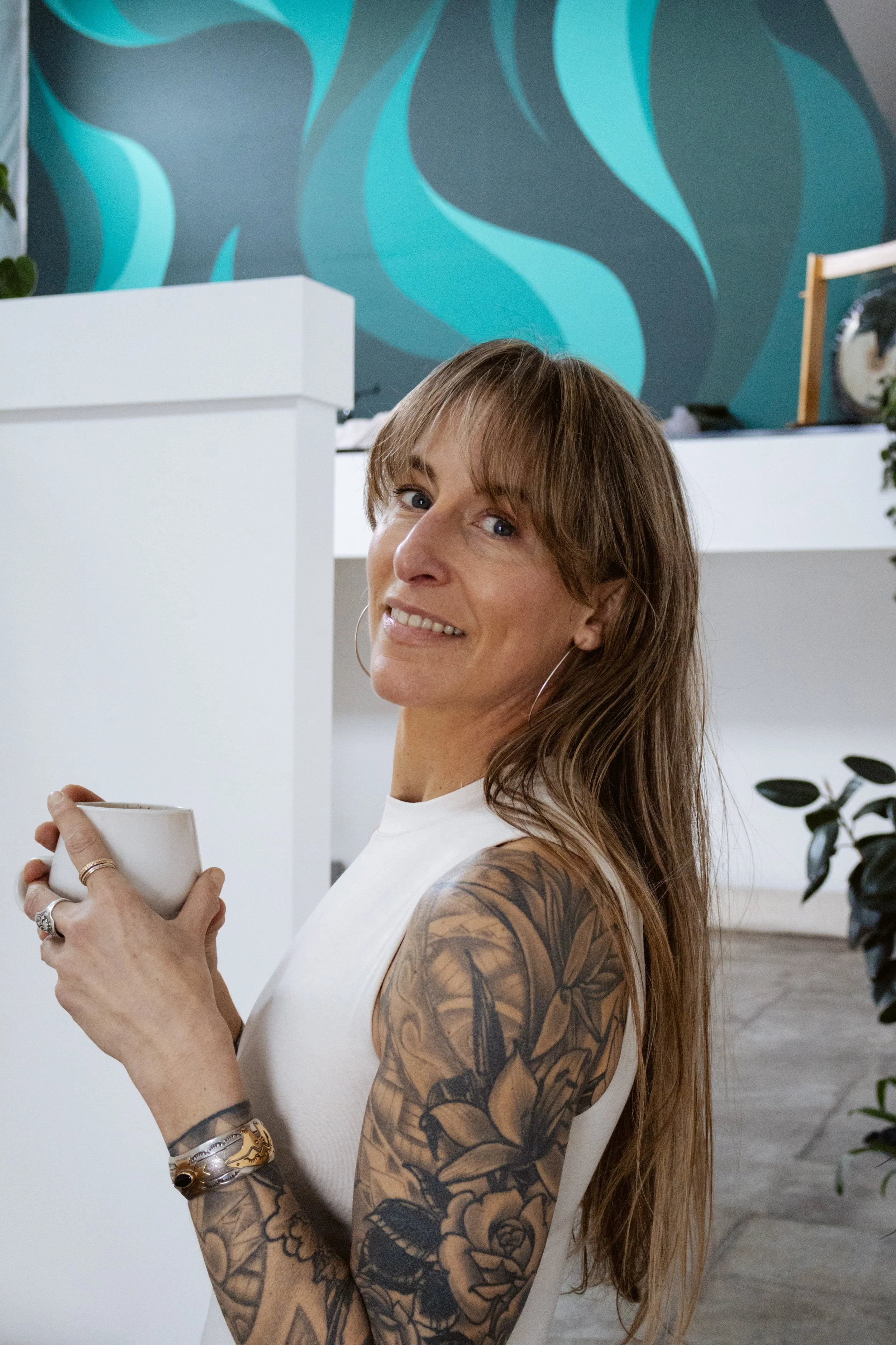 Smiling white woman physical therapist with long blonde hair wearing a heart-shaped pendant in front of a brown wall
