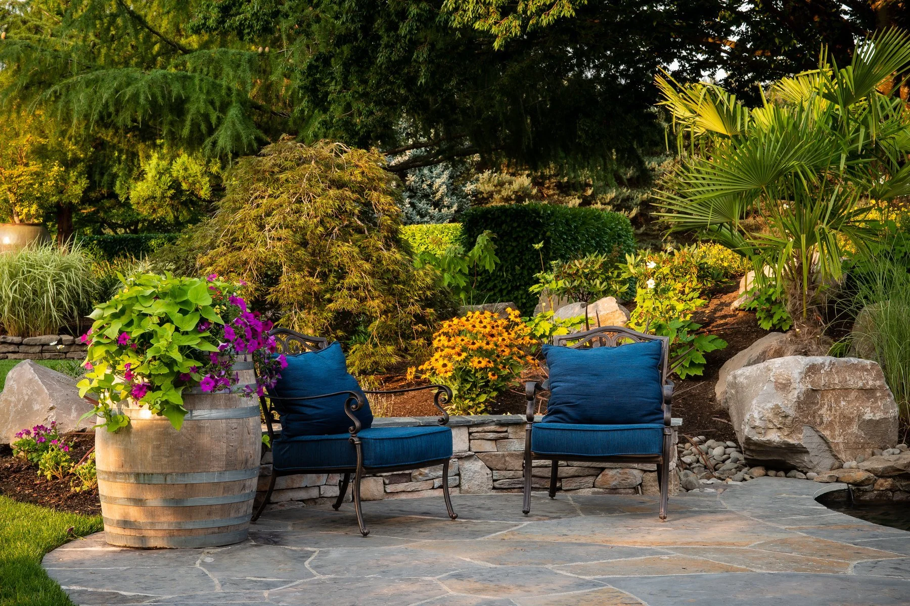 A cozy outdoor seating area in Tigard features blue armchairs and a rustic wine barrel planter on a flagstone patio.