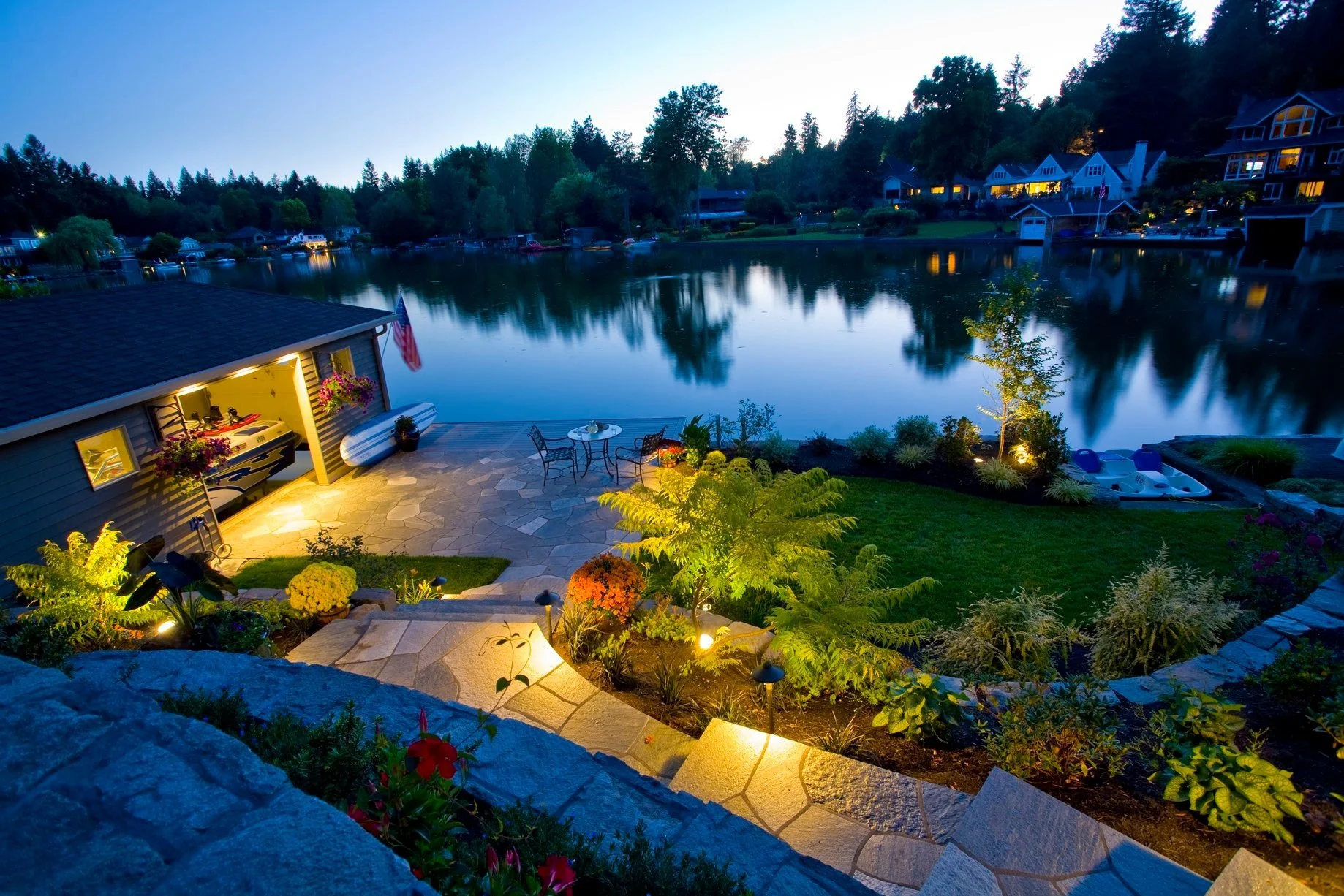 Stunning Tigard landscape design featuring illuminated stone steps leading down to a serene lakeside patio at dusk.