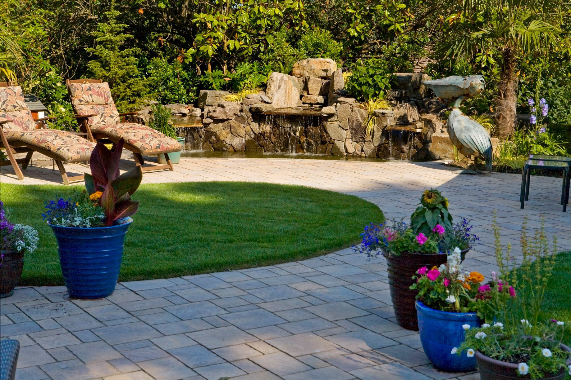A tranquil stone waterfall pours into a pond surrounded by a spacious paver patio in a Beaverton backyard.
