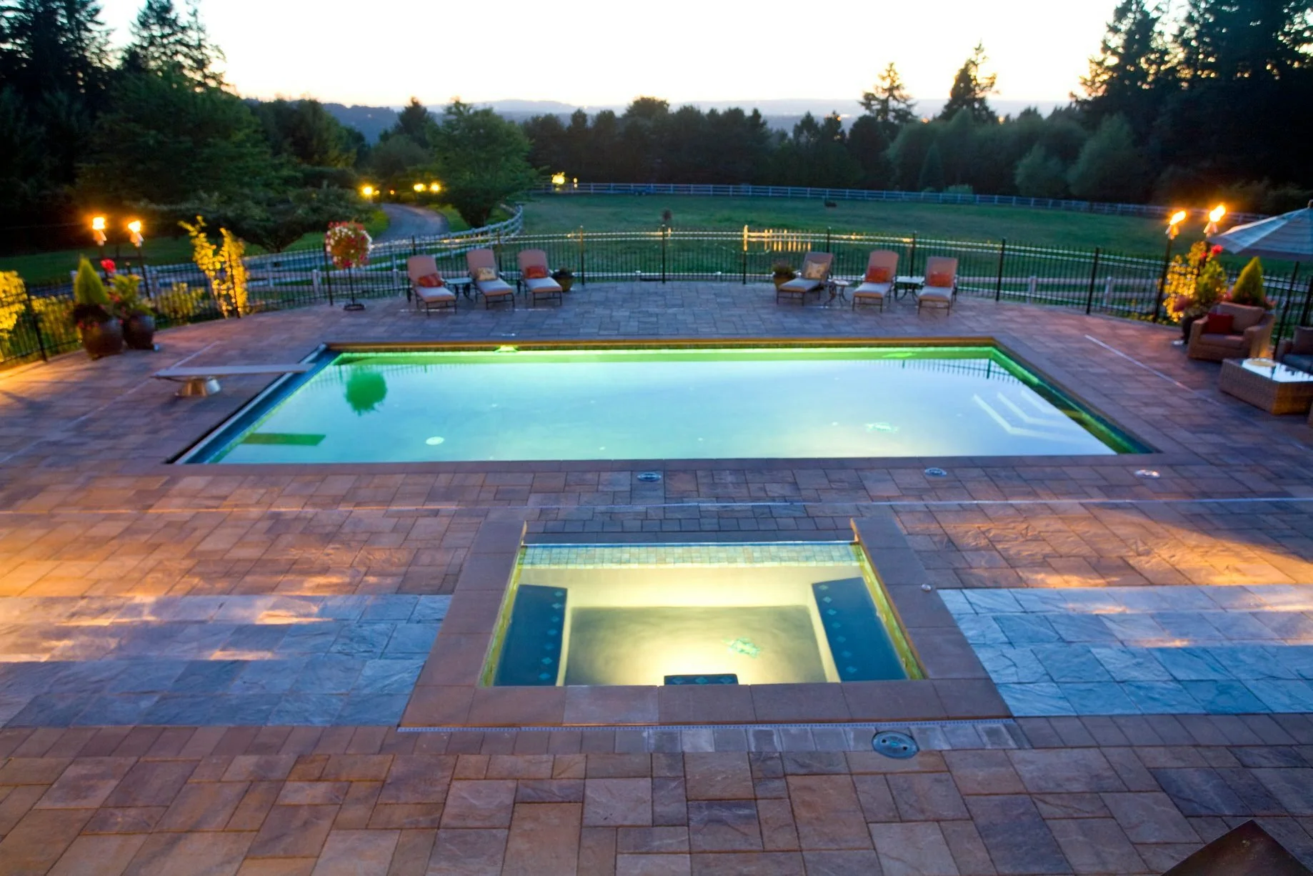 A sprawling paver pool deck overlooks a scenic Beaverton valley, perfectly illuminated by professional landscape lighting.