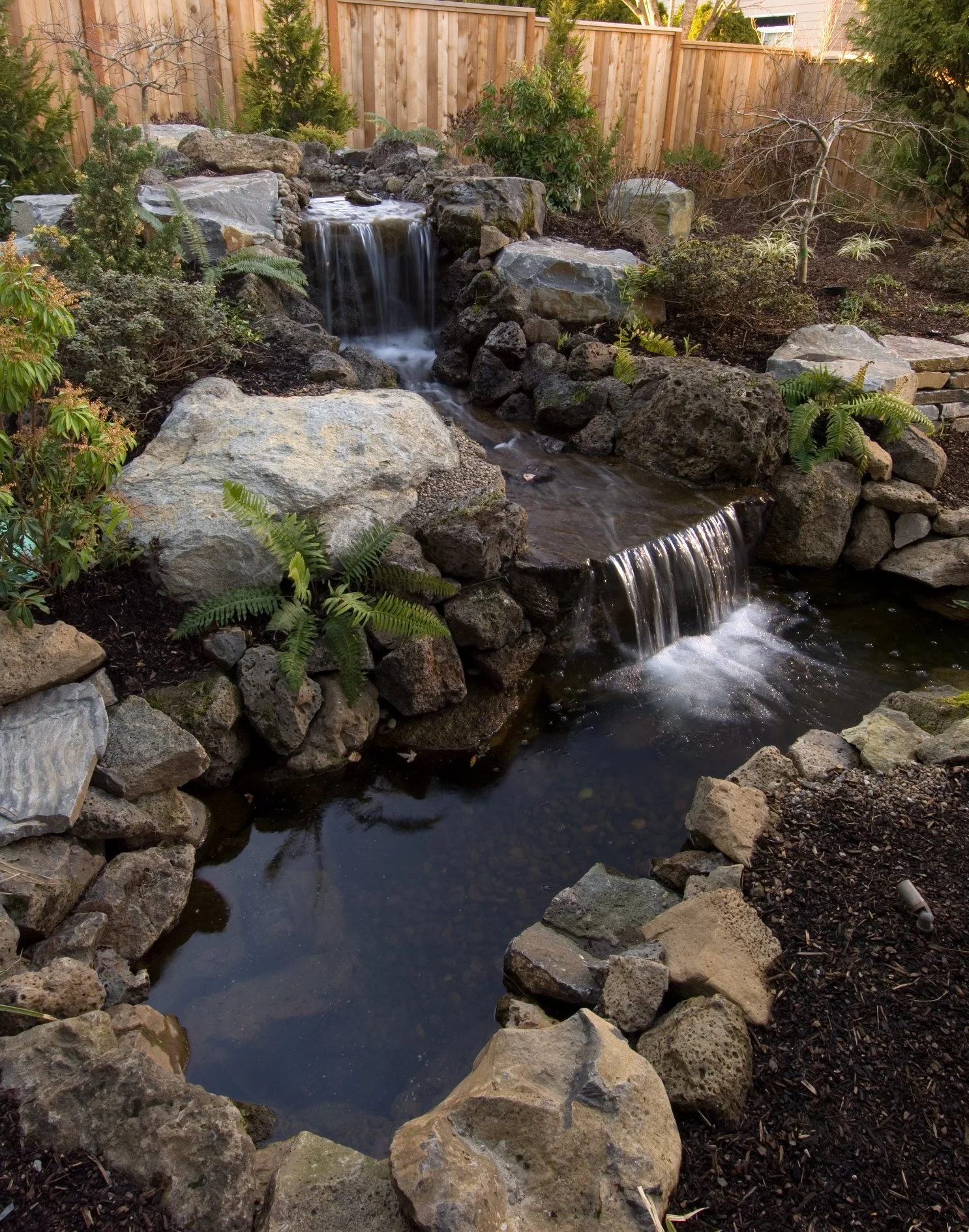 A multi-tiered natural rock waterfall flows into a tranquil pond within a private Tigard backyard.