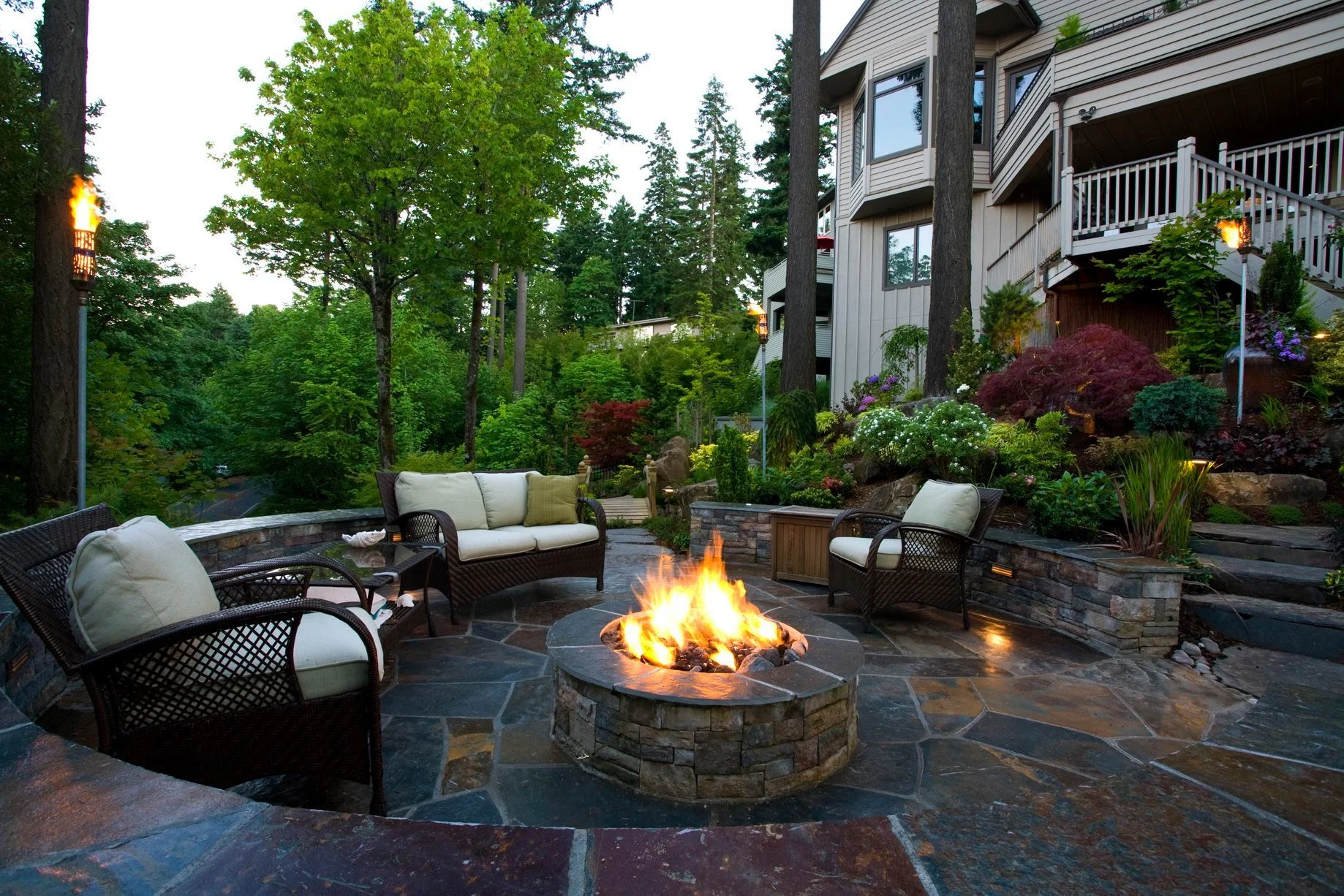 A luxury slate patio in Wilsonville features a stone fire pit, comfortable wicker seating, and tiki torch lighting for evening ambiance.