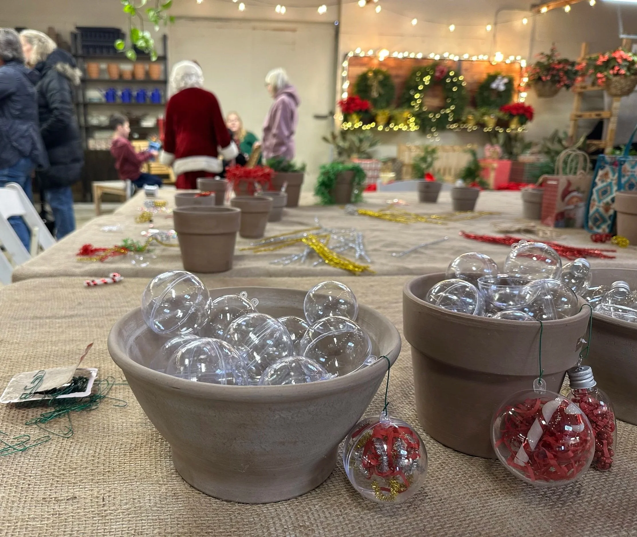 A table with clear plastic ornaments, some filled with red and silver decorations, and others empty, surrounded by small potted plants, with a festive holiday setting and people in the background.