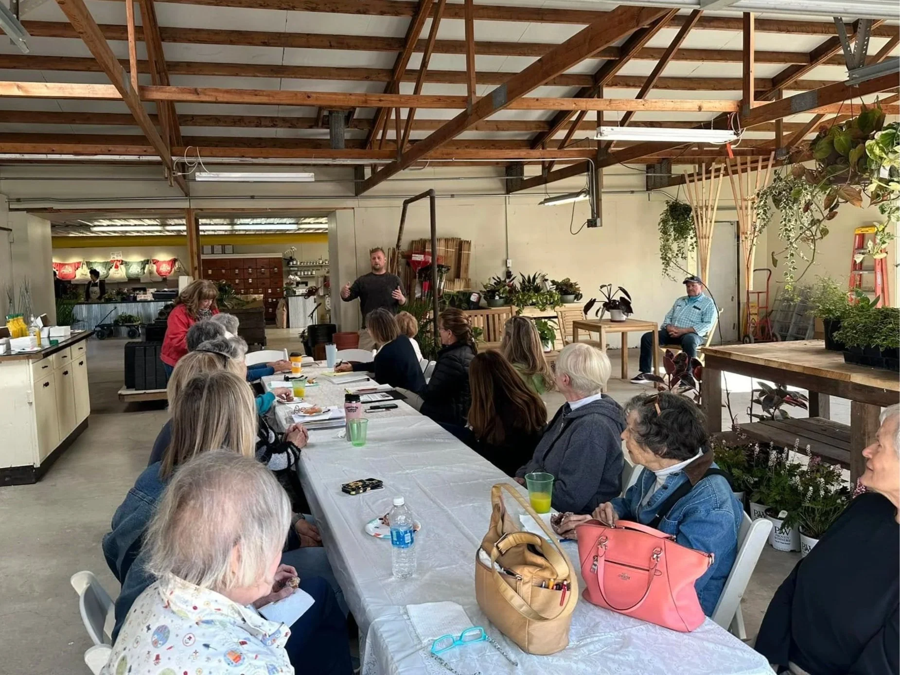 A group of people sitting at a long table in a workshop or classroom setting, listening to a speaker standing at the front. The room has exposed wooden beams, plants, and a casual, rustic atmosphere.