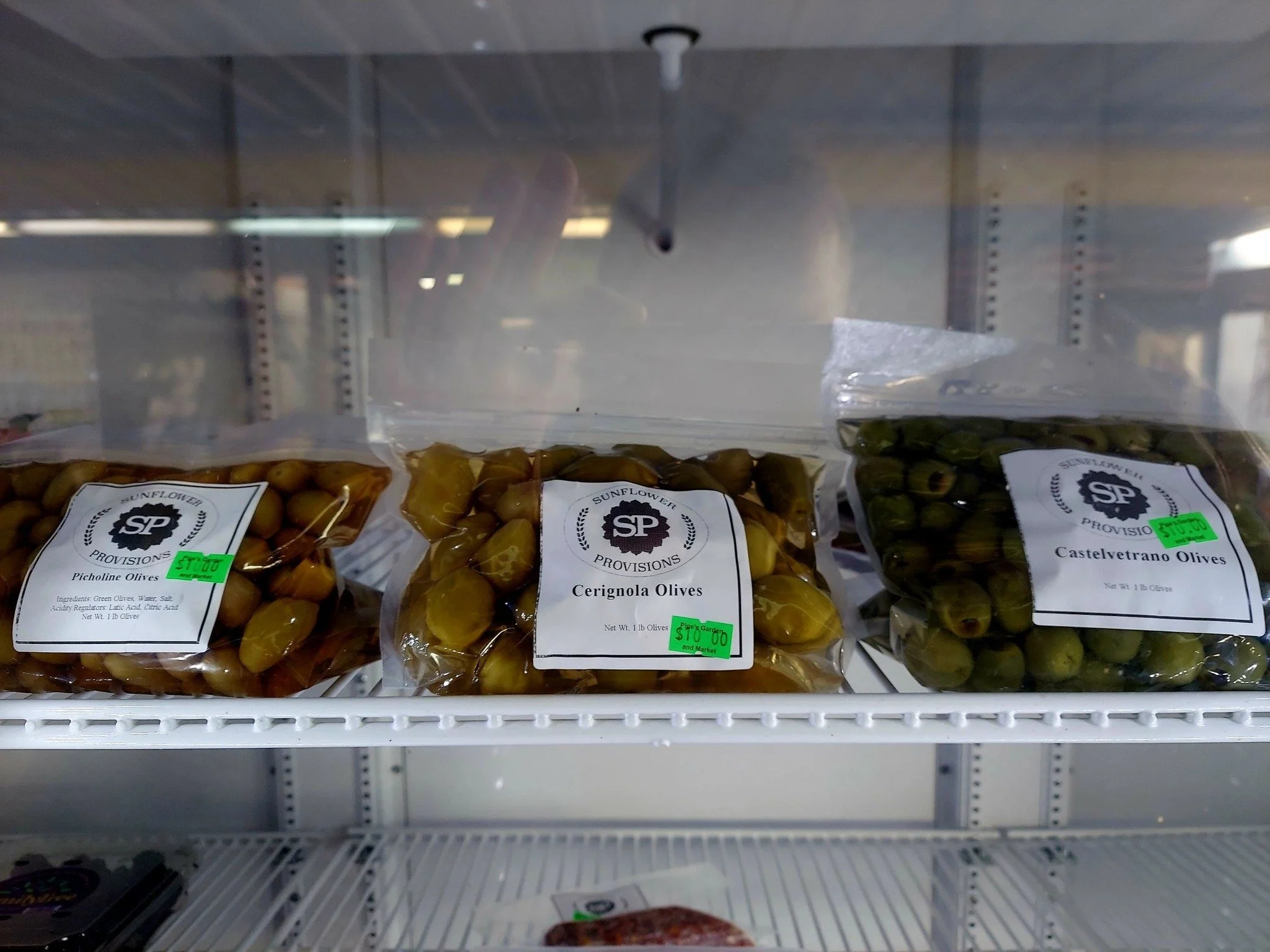 Bags of olives on a refrigerated shelf, including Piccoline Olives, Cerignola Olives, and Castelvetrano Olives, each labeled with green price stickers.