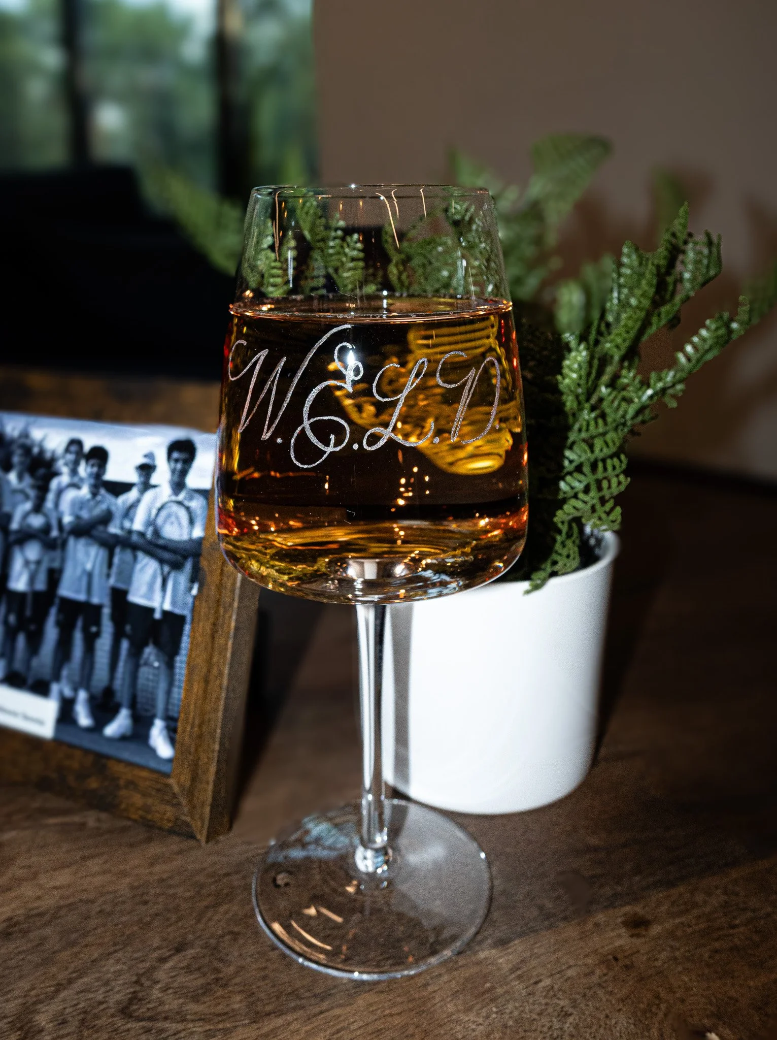 A wine glass filled with whiskey on a wooden table, with a black and white photo in a wooden frame and a potted plant in the background.