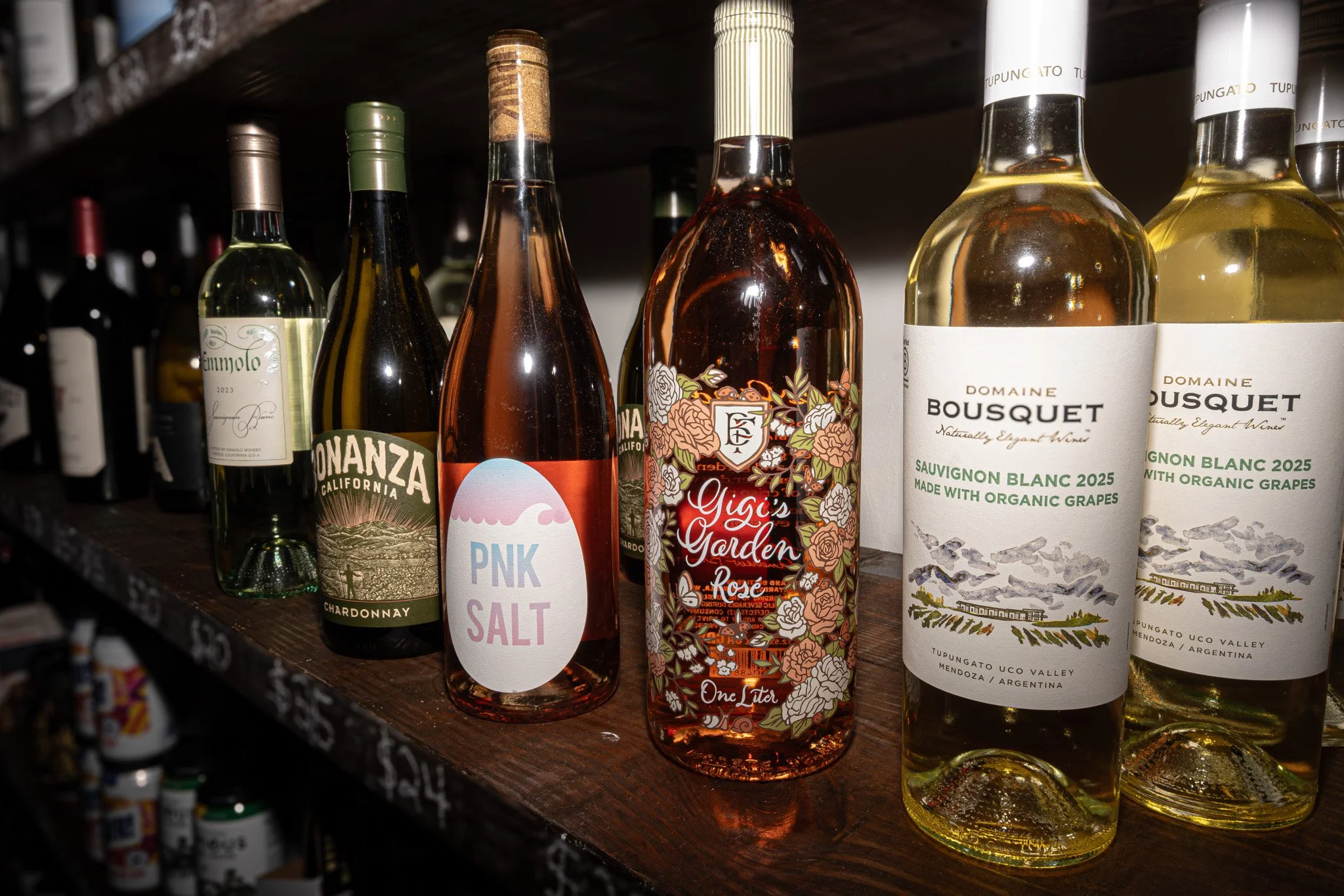 Line of various wine bottles on a wooden shelf, including Chardonnay, Sauvignon Blanc, and rosé, with some labels visible.