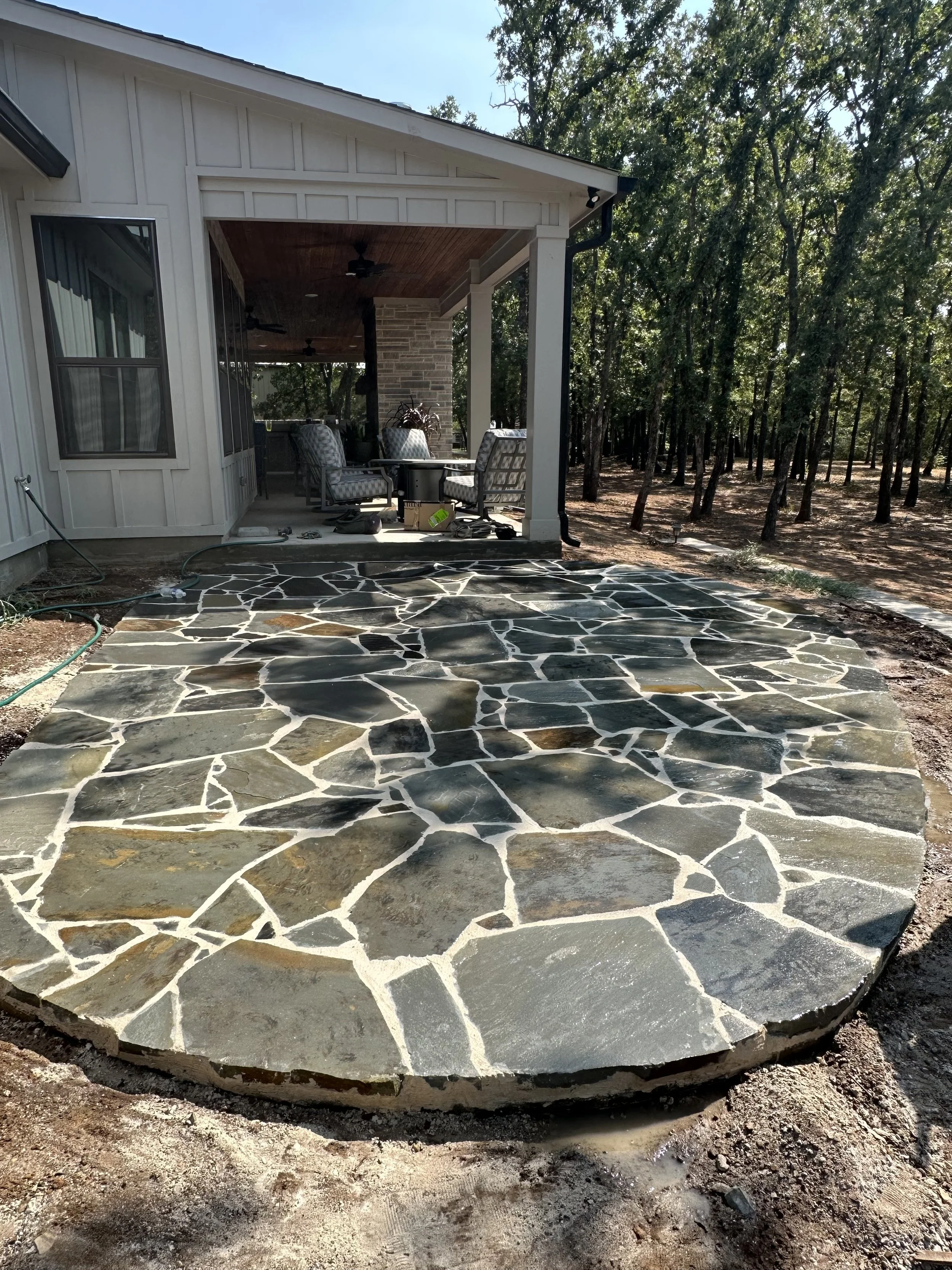 View of a new stone patio with irregular dark and light colored stones, attached to the porch of a house with outdoor furniture, in a wooded backyard.