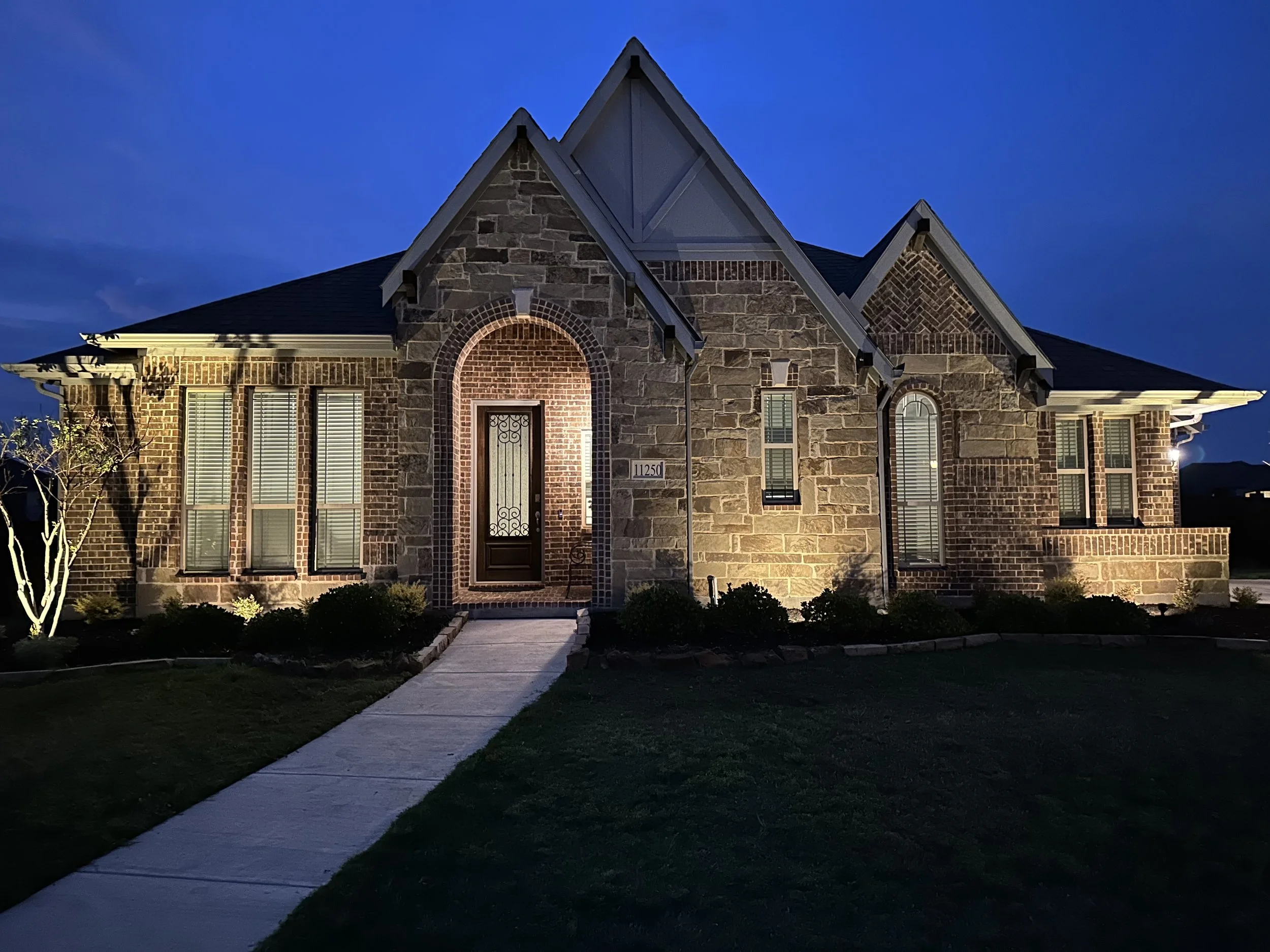 Front view of a brick house illuminated at night, with a sidewalk leading up to the entrance and some small bushes and trees in the yard.