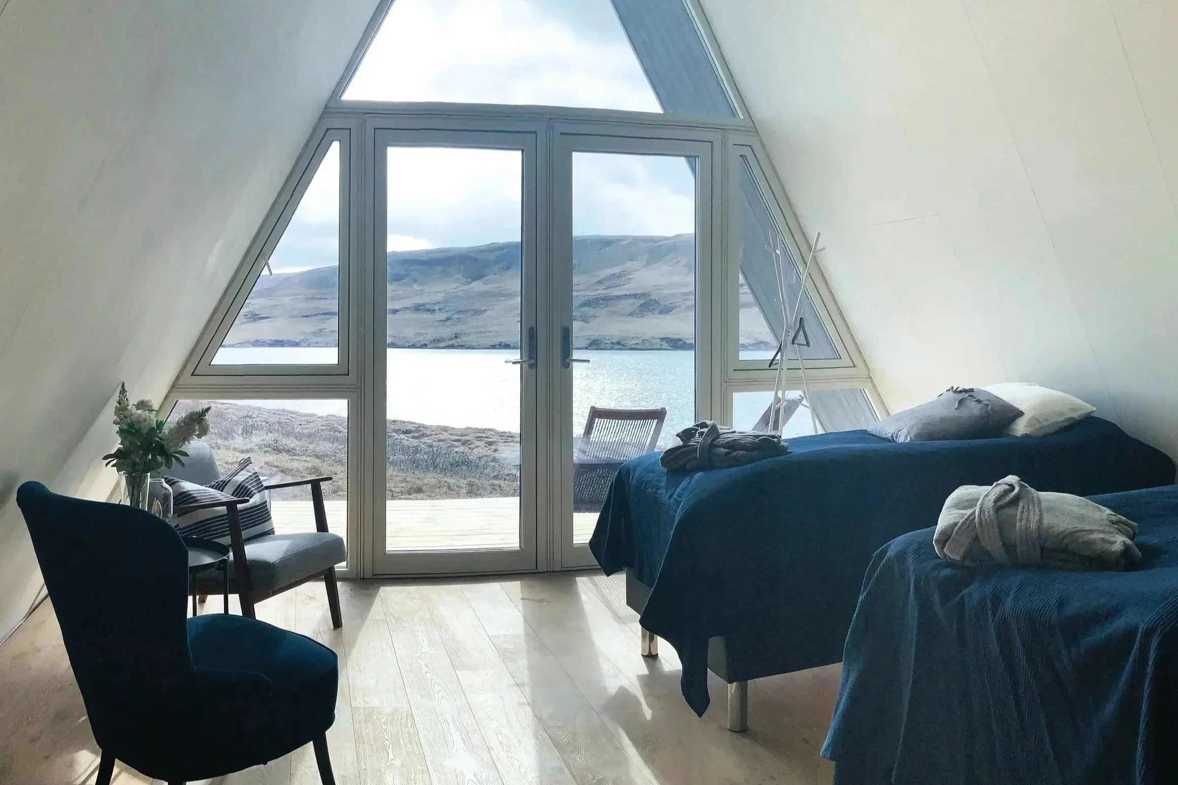 Interior of a cozy room with two beds covered in dark blue blankets, notions of rolled towels, a chair with a cushion, a small table with a vase of flowers, and large windows opening to a scenic lake and mountain view.