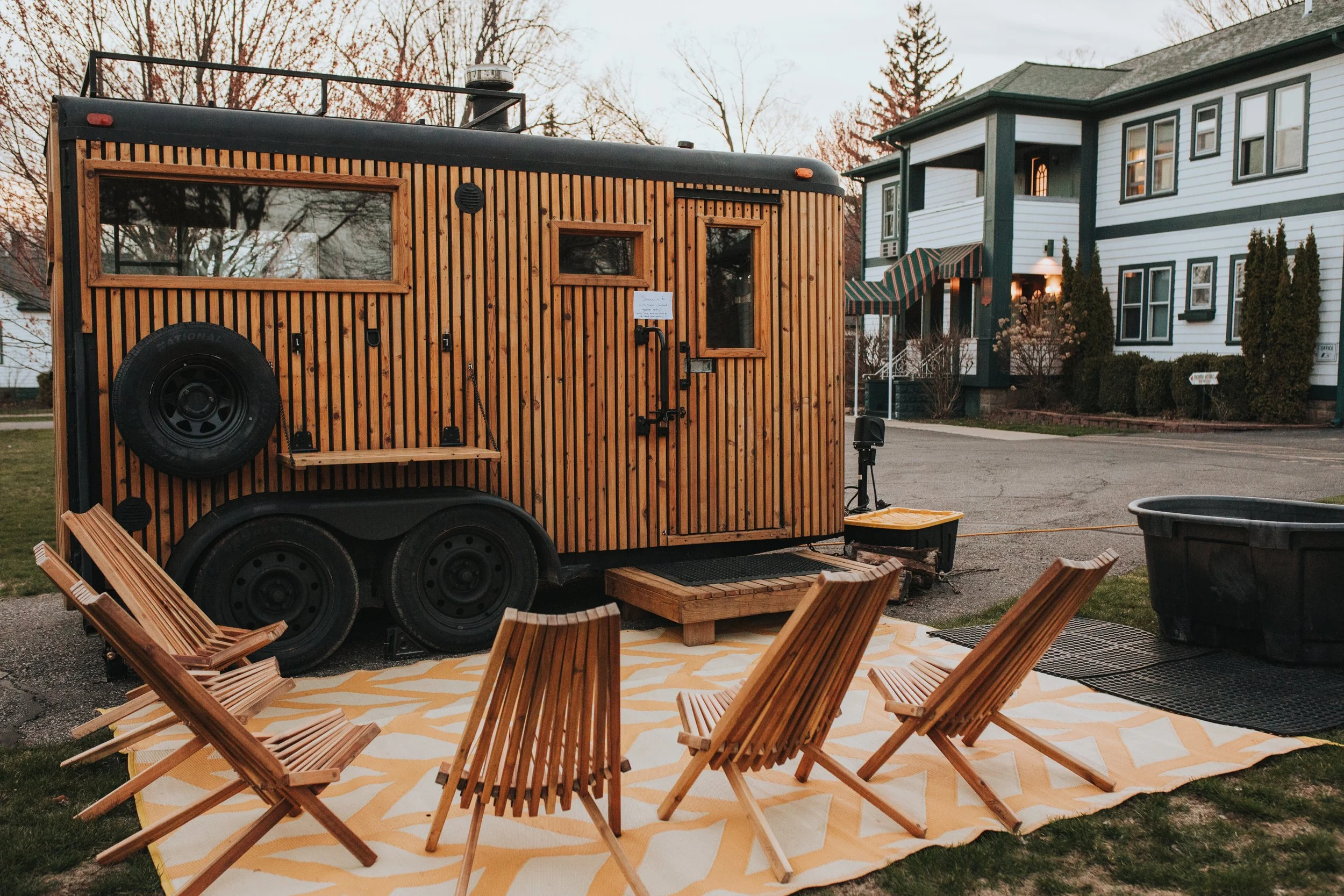 A tiny house on wheels with vertical wooden slat siding, black window frames, and a black ladder leading to the roof, parked outdoors.