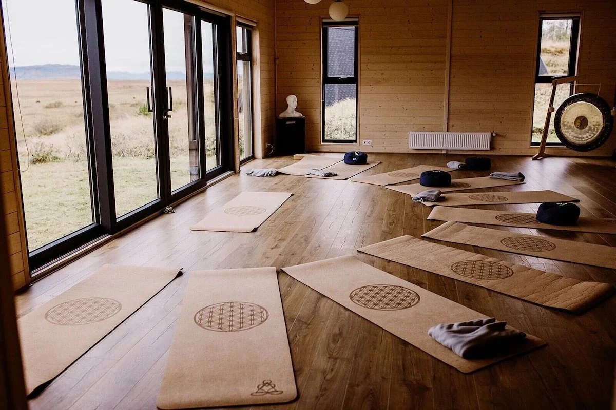 Yoga mats arranged on a wooden floor in a room with large windows overlooking a natural landscape, with meditation cushions and a gong in the corner.