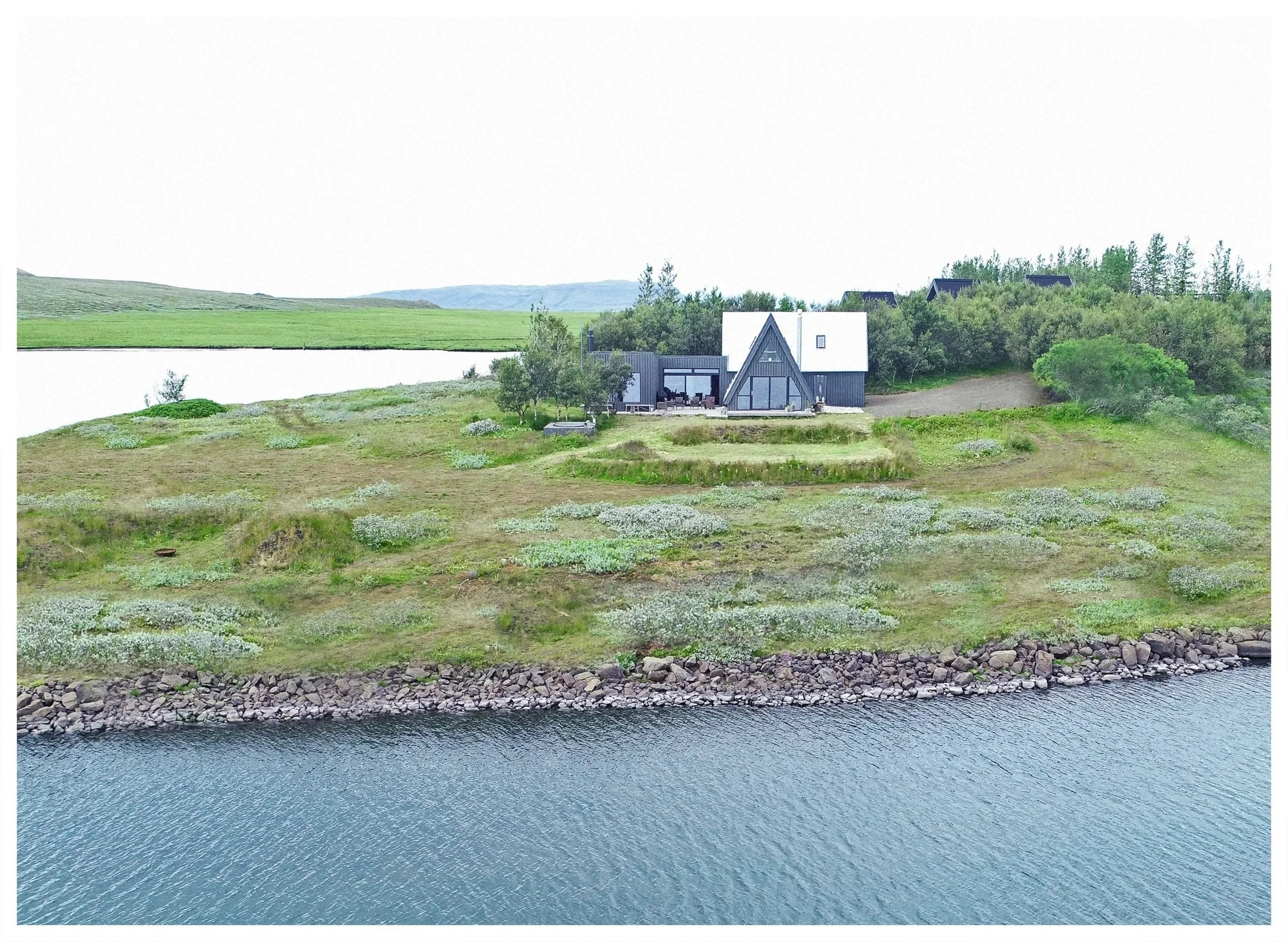 A modern house with black exterior and large windows situated on a green hillside by a body of water, with trees and open land in the background.