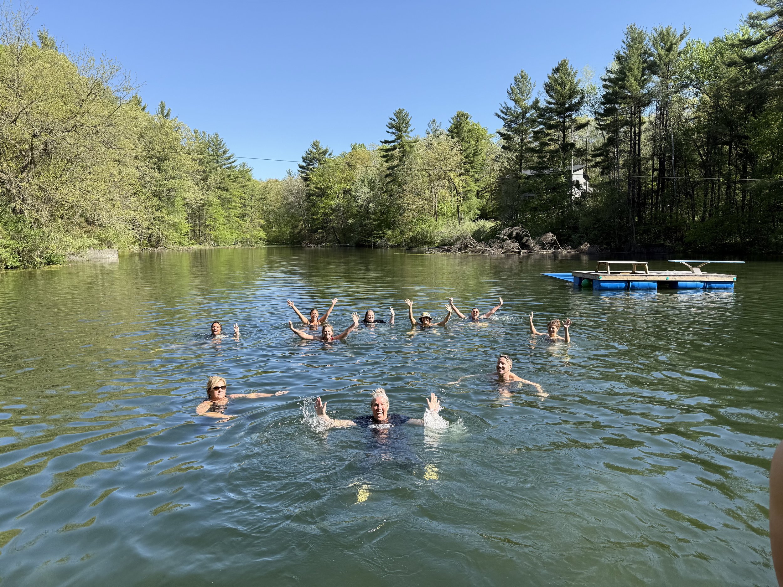 People swimming and enjoying themselves in a natural outdoor body of water, surrounded by lush green trees under a clear blue sky.