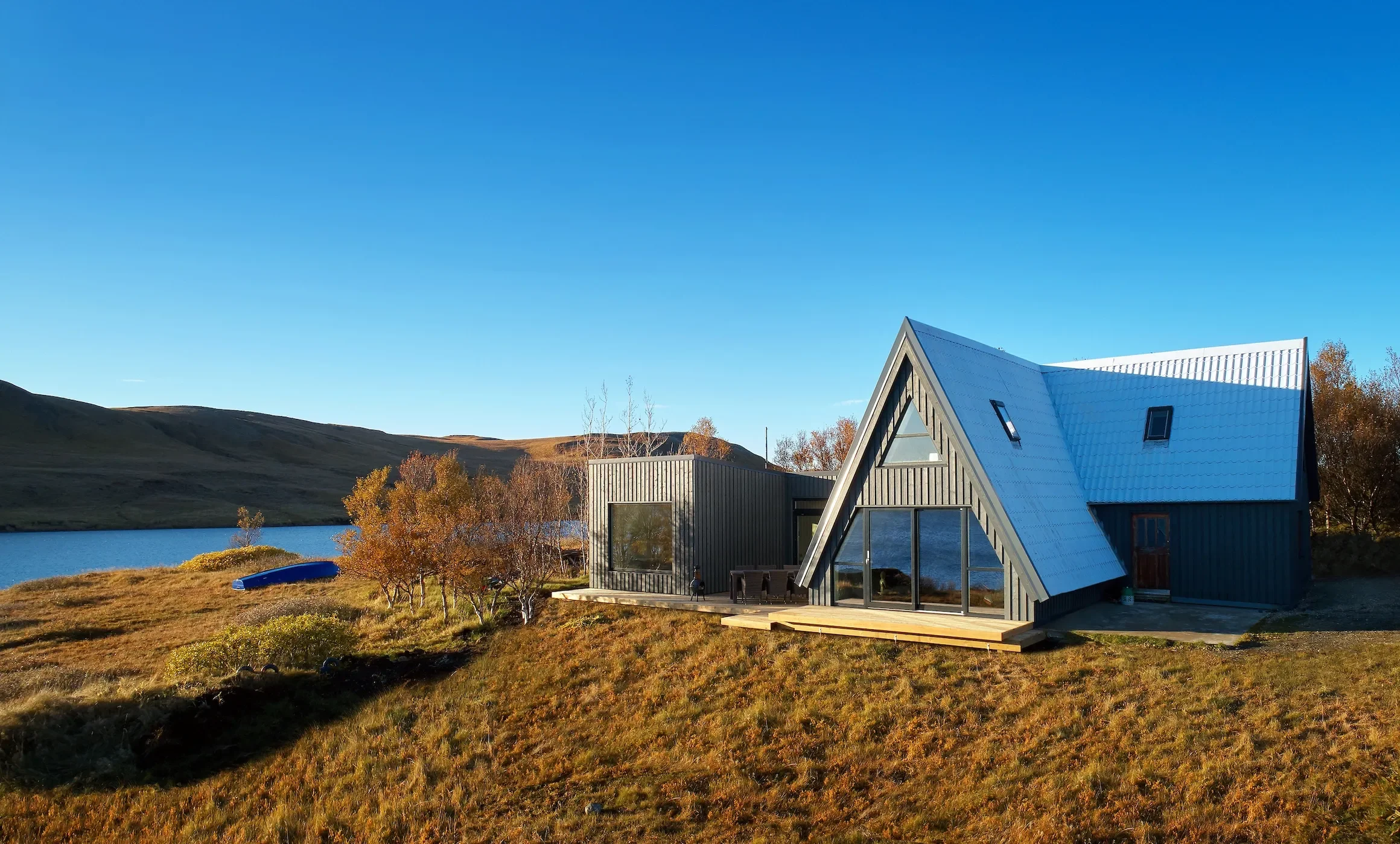 Modern house with a steeply pitched roof on a grassy landscape with trees, near a lake, under a clear blue sky.