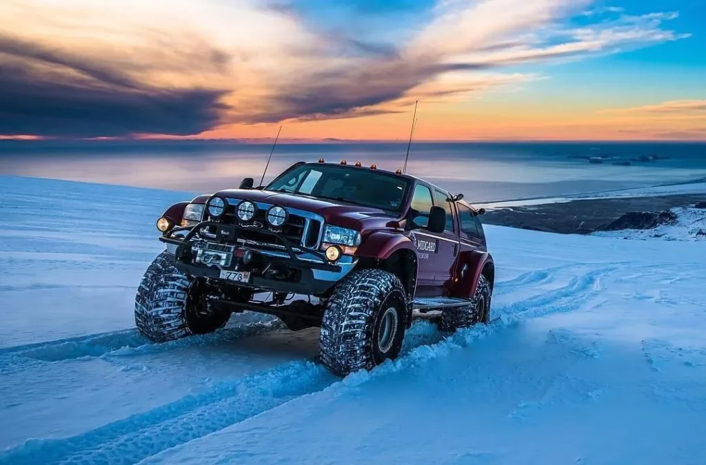 A red off-road vehicle driving on a snow-covered landscape with a sunset sky in the background.