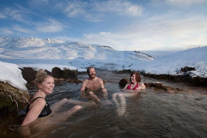 Three people enjoying a hot spring outdoors in a snowy landscape with mountains and blue sky in the background.