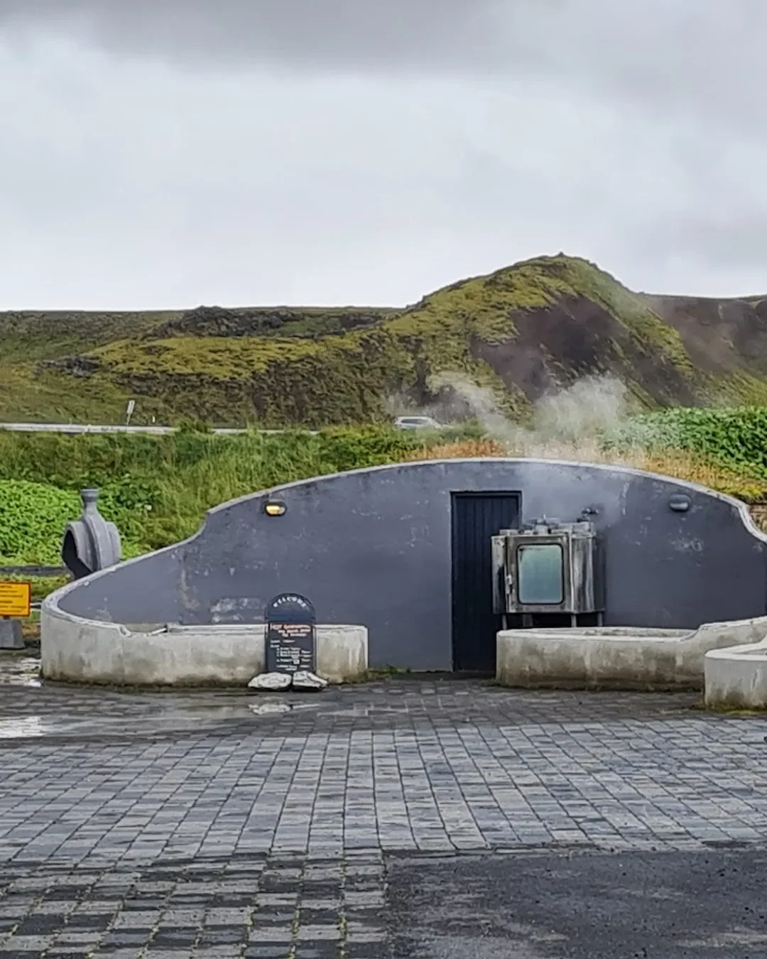 A geothermal hot spring with steaming water, surrounded by a paved area and a black fence, with green hills and cloudy sky in the background.