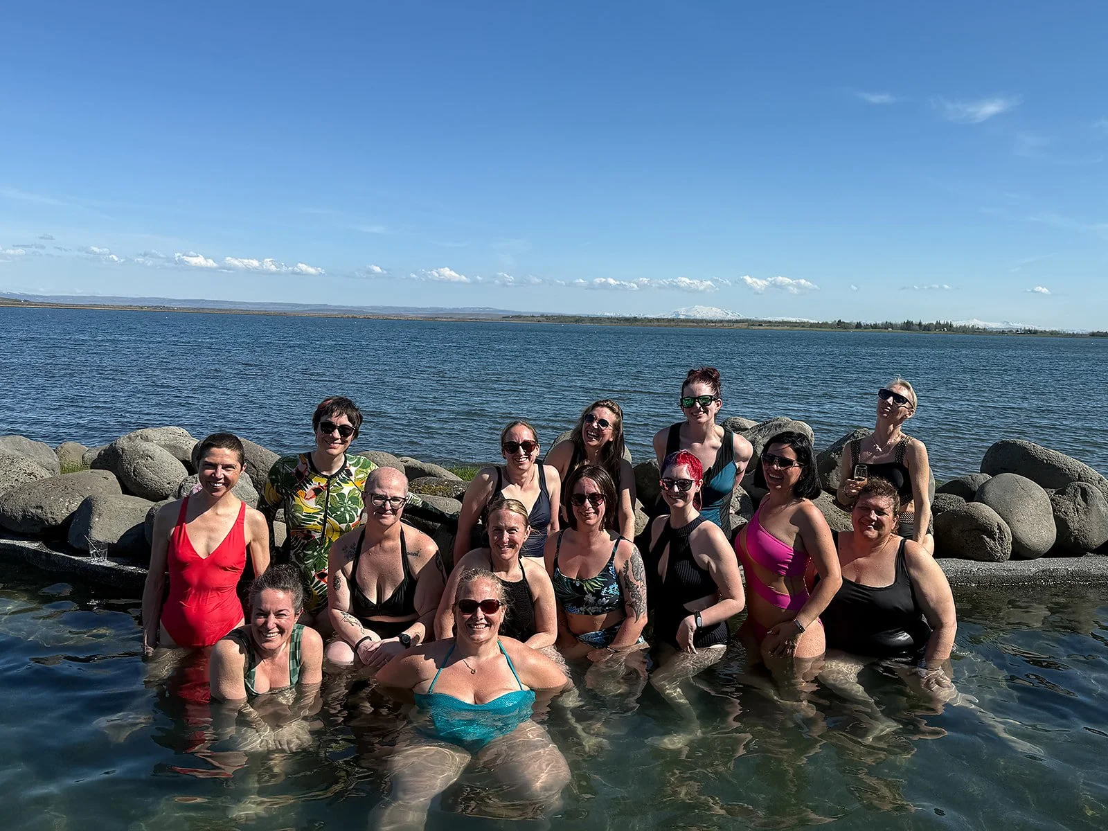 Group of people in swimwear standing in water by rocks, with a large body of water and sky in the background.