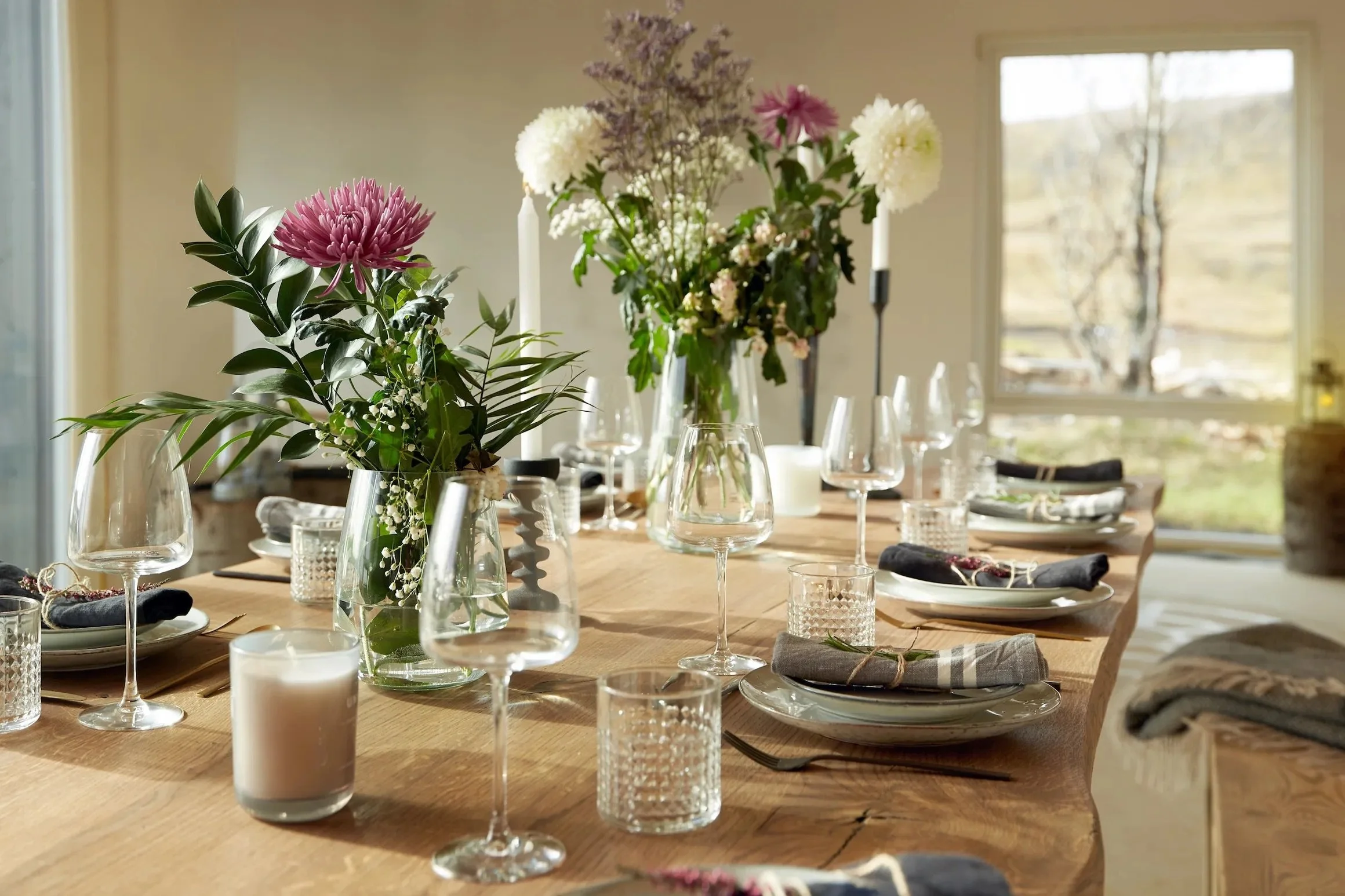 A well-set dining table decorated with floral centerpieces, wine glasses, candles, and plates, illuminated by natural light from large windows showing an outdoor view.
