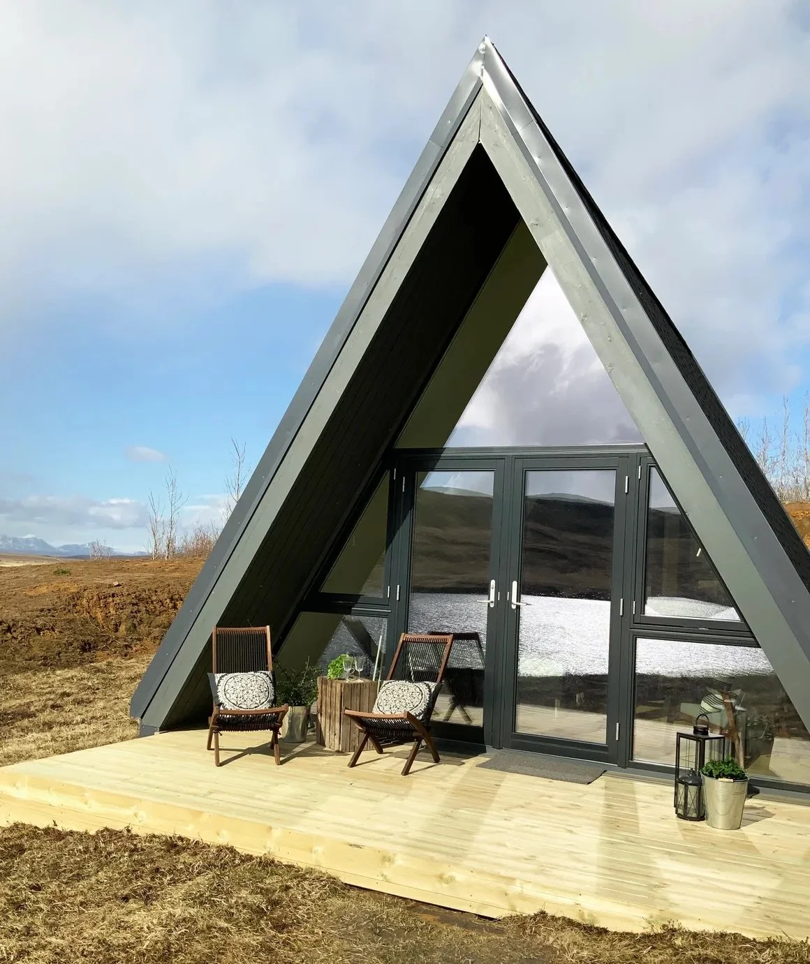 Aerial A-frame tiny house with glass doors, outdoor wooden deck, two chairs, and potted plants in a natural landscape.