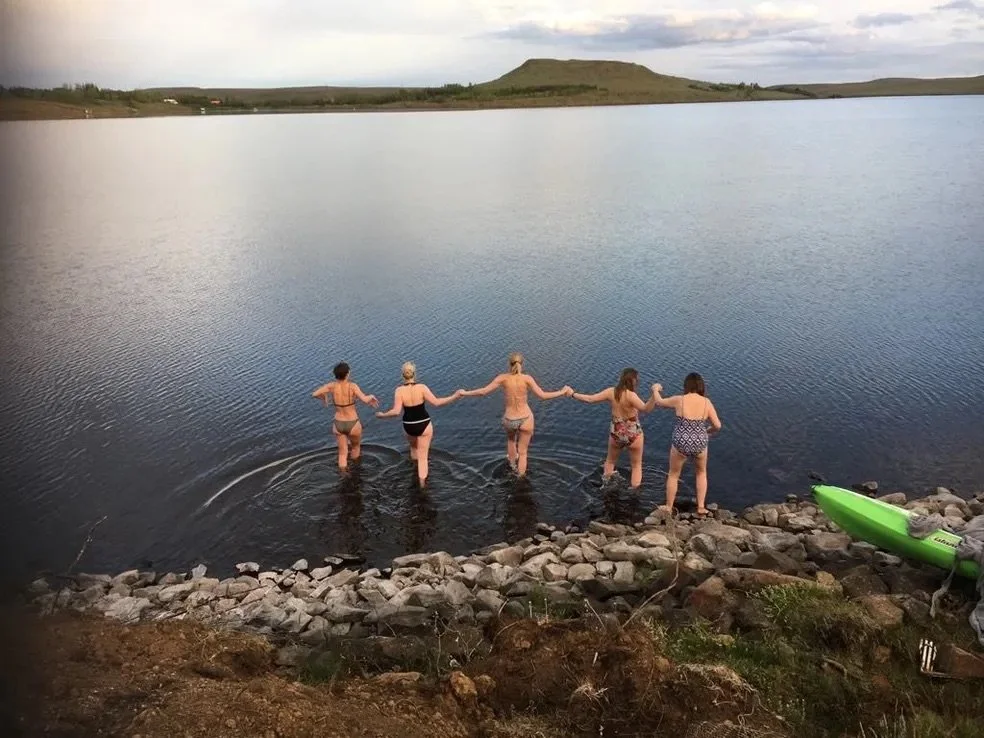 Five women in swimsuits holding hands, walking into a lake from a rocky shore during daytime.