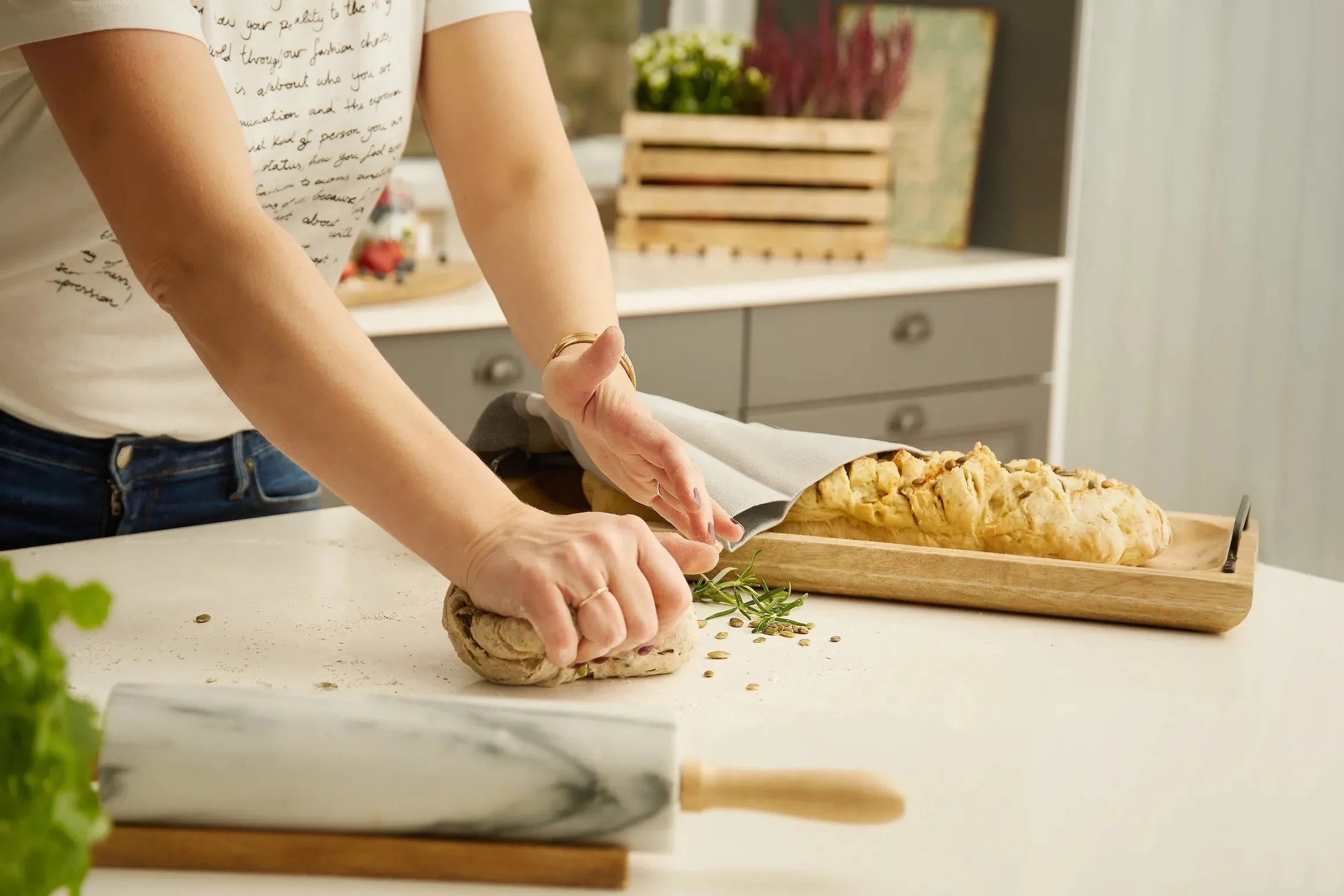 Person kneading dough on a kitchen countertop with fresh herbs, with a loaf of bread on a tray nearby.