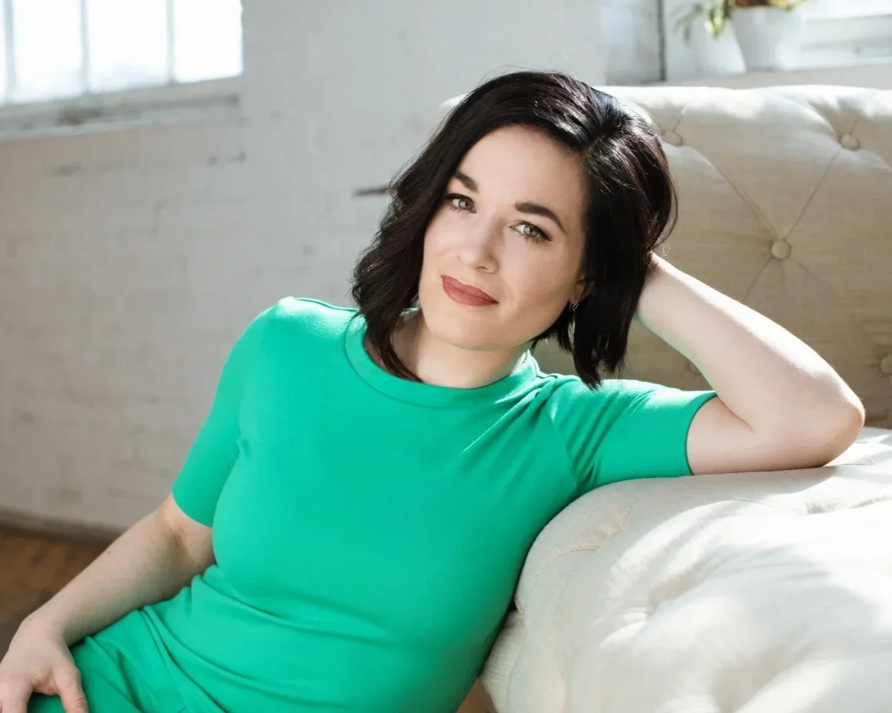 A woman with short dark brown hair, wearing a bright green shirt, sitting on a sofa in a bright room with white brick walls and large windows, resting her head on her hand and smiling at the camera.