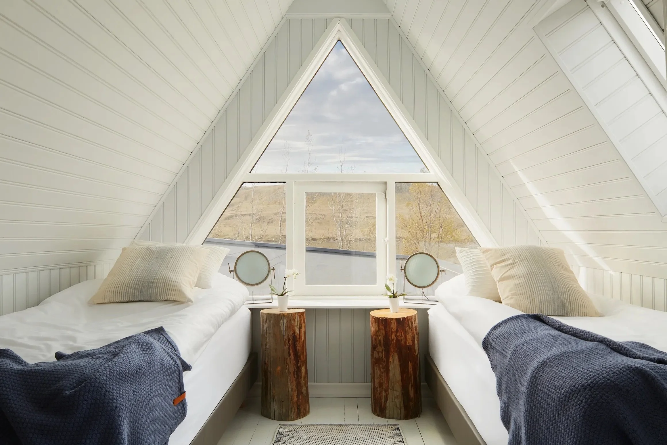 A cozy attic bedroom with two single beds, each with beige pillows and navy blankets. The beds are separated by two wooden stump bedside tables. A large triangular window at the end of the room offers a view of the outdoors with trees and cloudy sky, and small round mirrors are placed on the window sill.