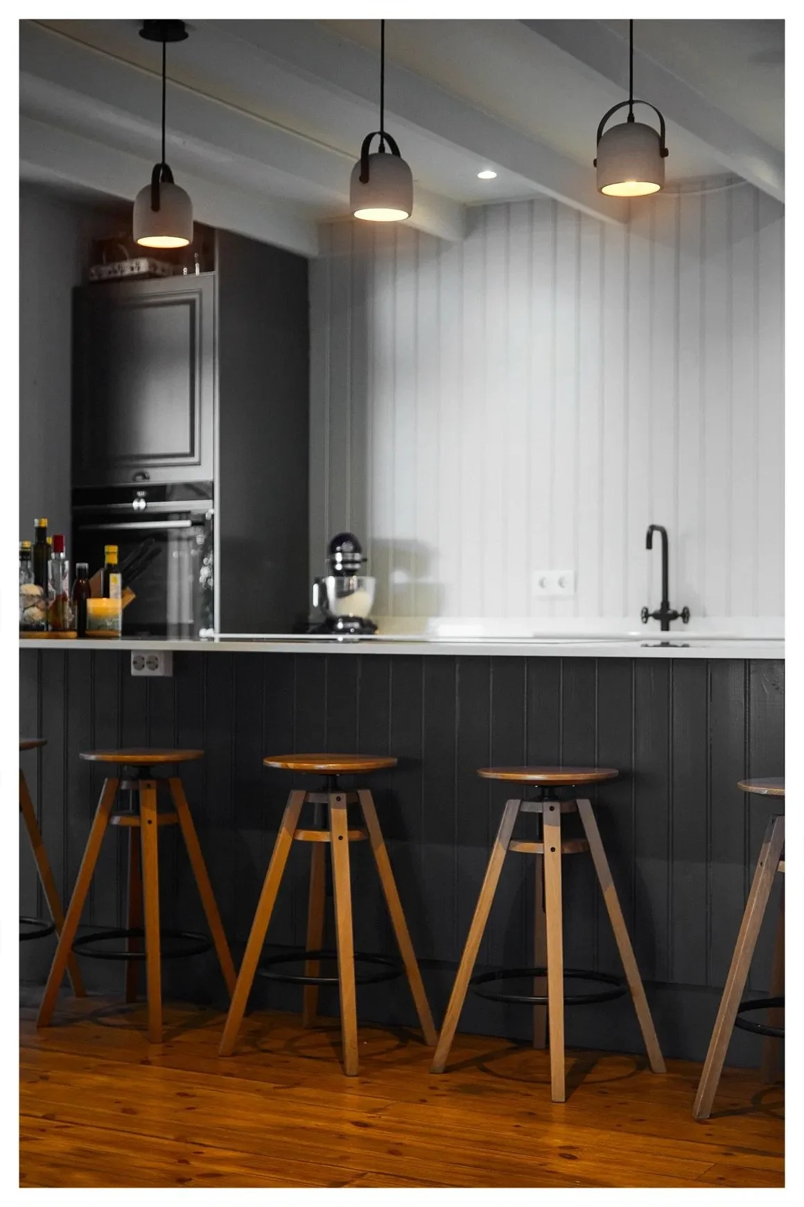 Interior of a modern kitchen bar area with four wooden stools, black and white cabinetry, pendant lights, and a wooden floor.