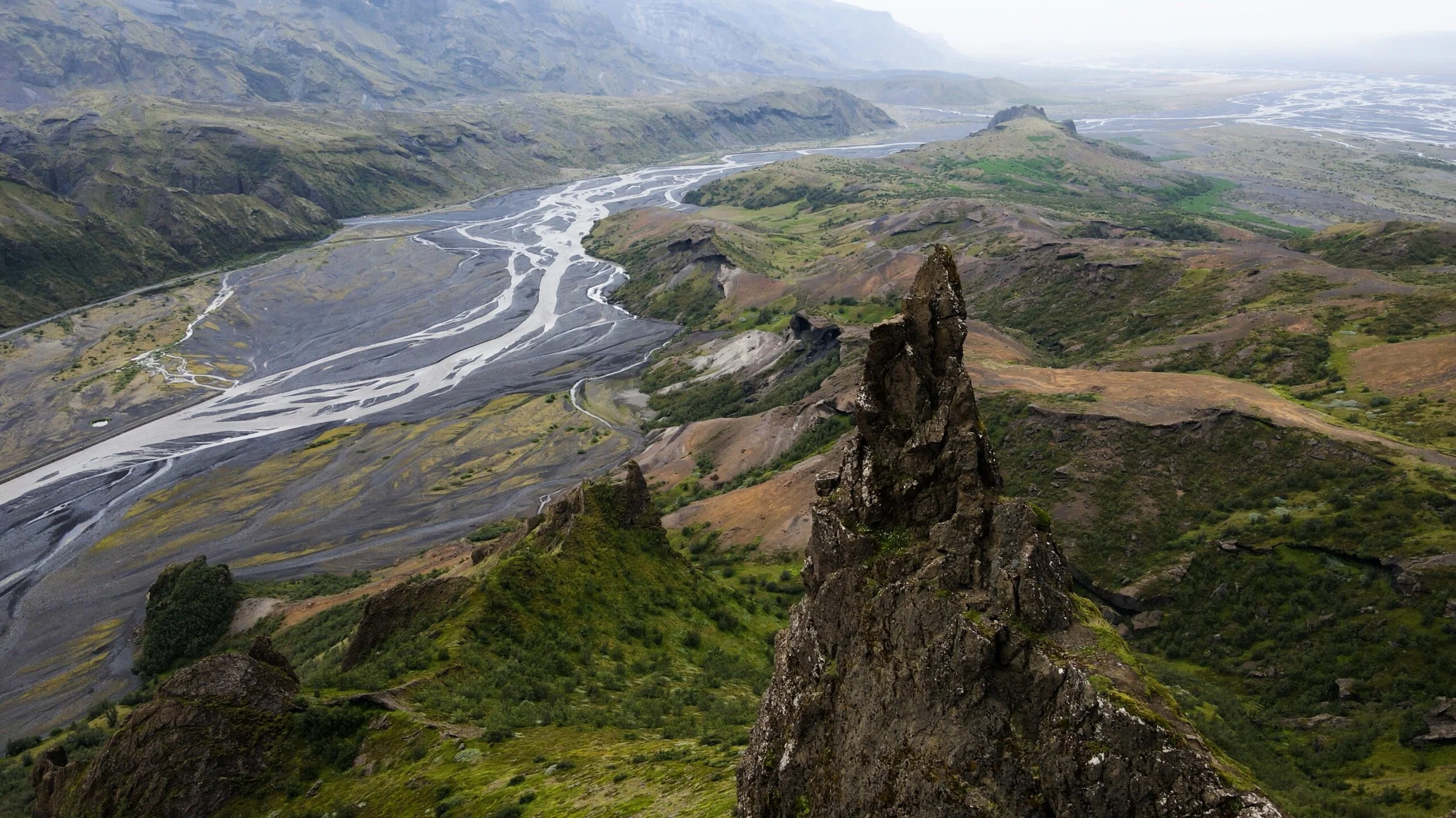 Aerial view of a valley with braided river channels, green hills, and rocky formations under a cloudy sky.