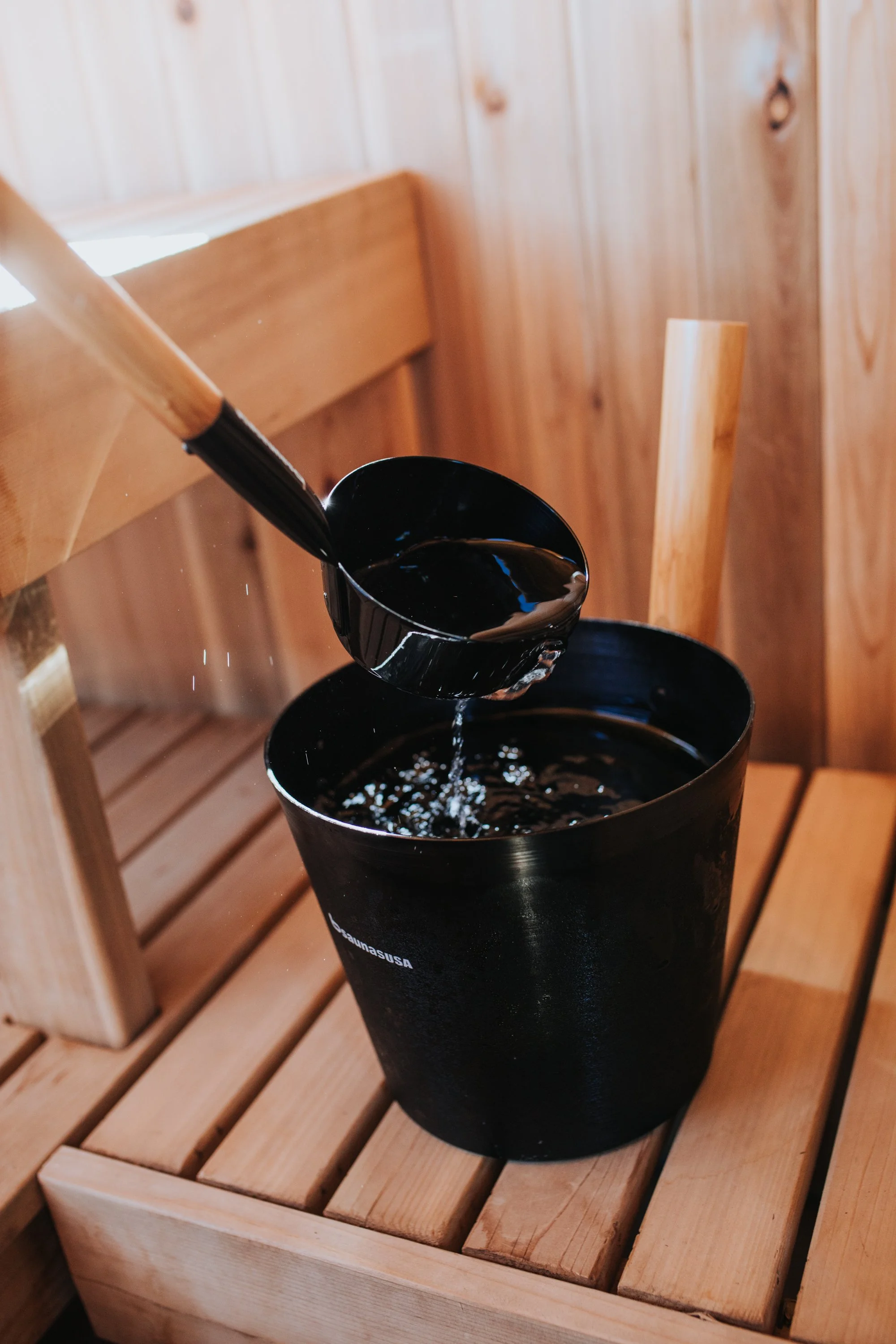 A black bucket with a metal ladle inside, placed on a blue cloth on a wooden surface, with three bottles labeled 'Rento' featuring labeled orange, green, and amber, against a wooden wall background.
