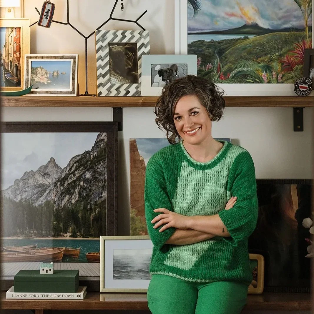 A woman with short curly hair wearing a green sweater and pants, smiling while sitting in front of a gallery wall filled with framed artwork and photographs, including landscapes and mountain scenes.
