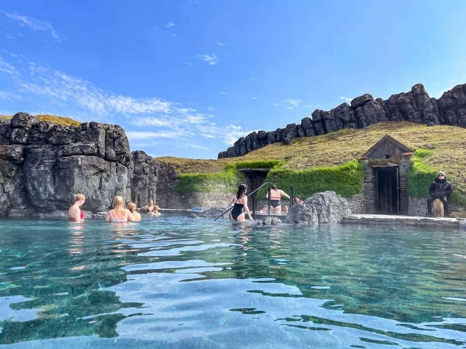People enjoying a geothermal bathing pool with rocky surroundings and a small wooden structure, under a partly cloudy blue sky.