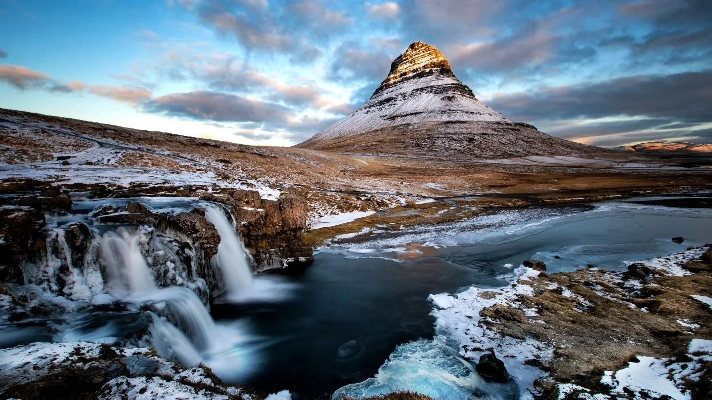 Snow-capped mountain with a waterfall, frozen lake, and cloudy sky.