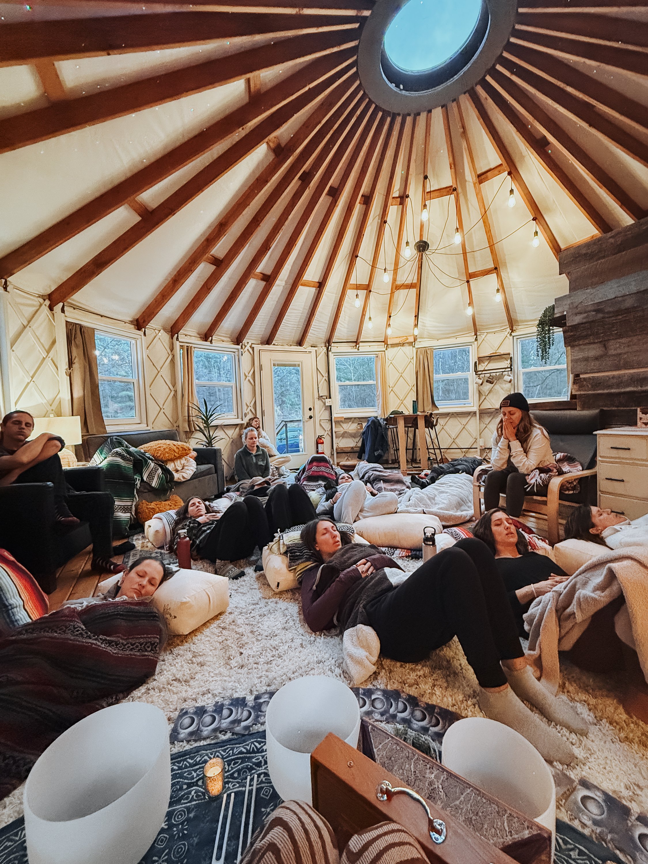 People relaxing and lying on cushions and blankets inside a circular yurt with wooden beams and windows, a modern chandelier hanging from the ceiling.