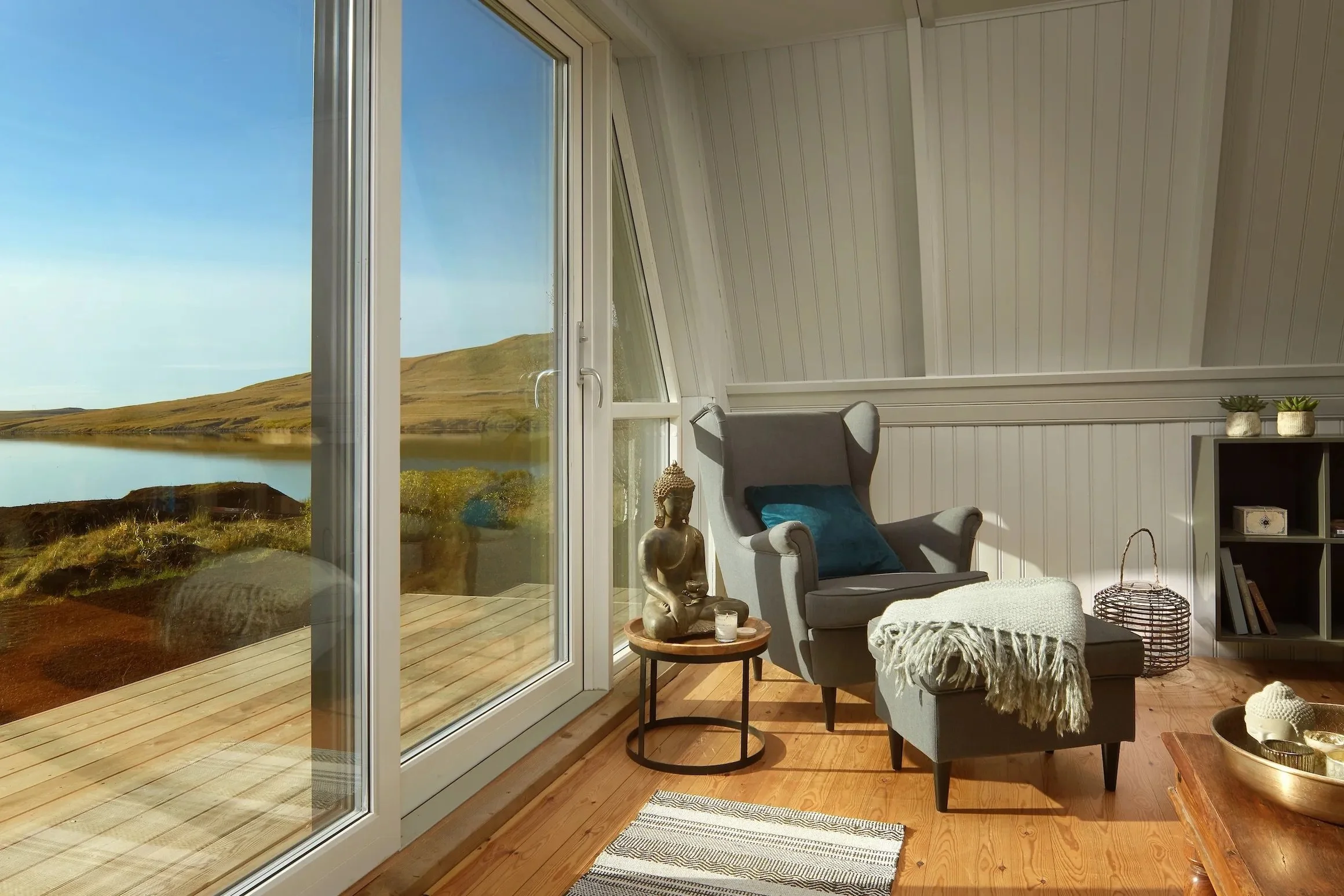 Sunlit room with a large window showing a scenic view of a lake and hills. Inside, a gray armchair with a blue pillow, a small side table with a Buddha statue, a candle, and a throw blanket. There is a wooden floor, striped rug, and a bookshelf with 