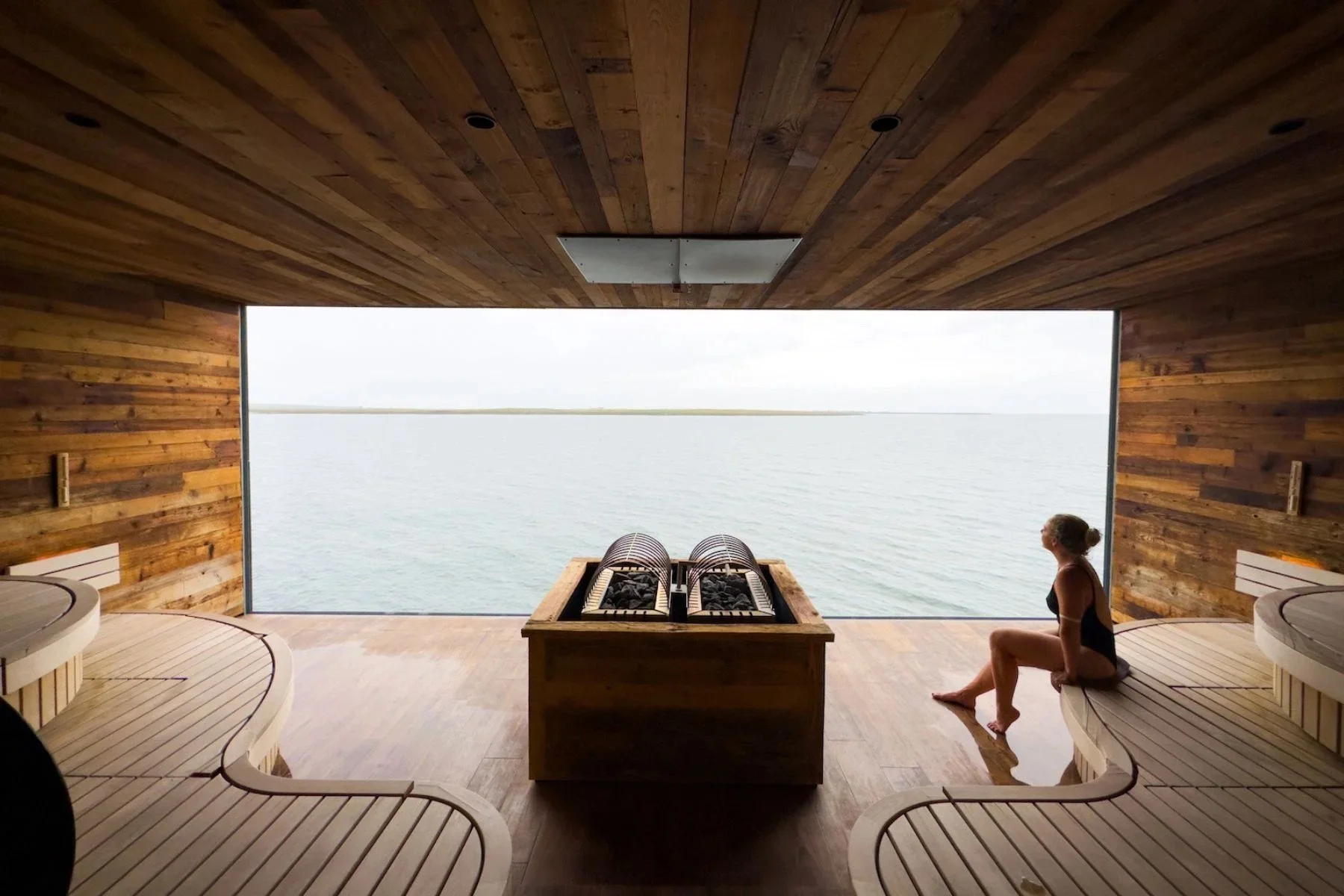A woman sitting on a wooden bench inside a sauna room, looking out through a large open window at a body of water and distant shoreline. The room has wooden walls and ceiling, with built-in seating and a heater in the center.